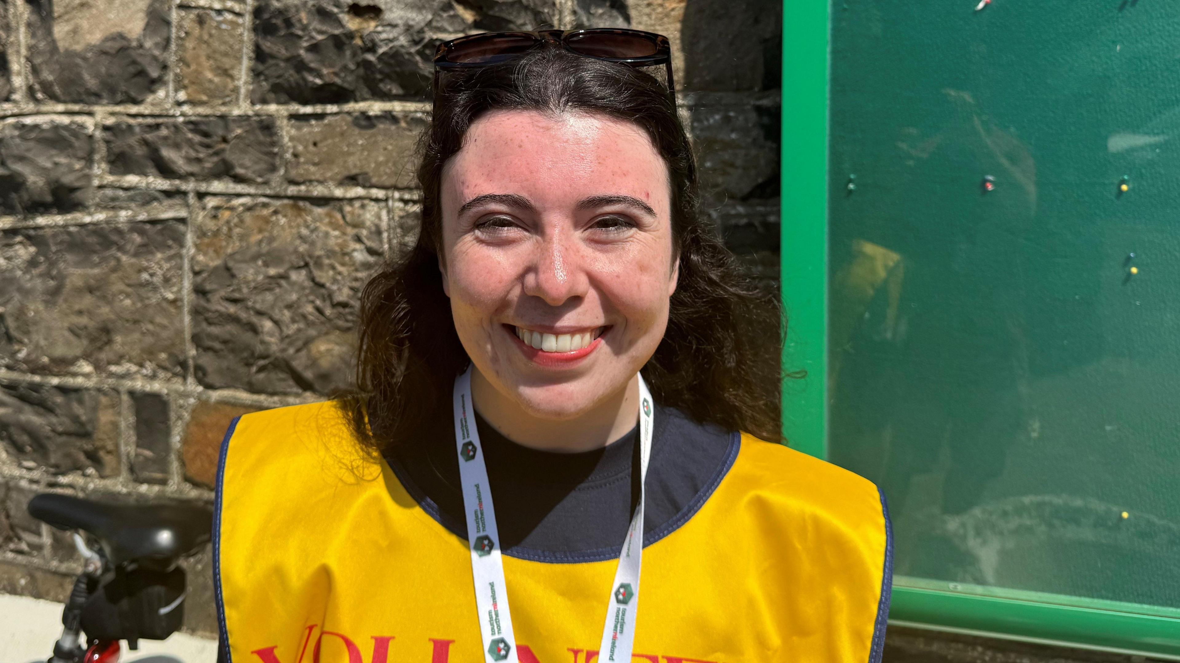 Keely is smiling. She has brown hair. She's wearing a yellow bib that says volunteer. She's outside.