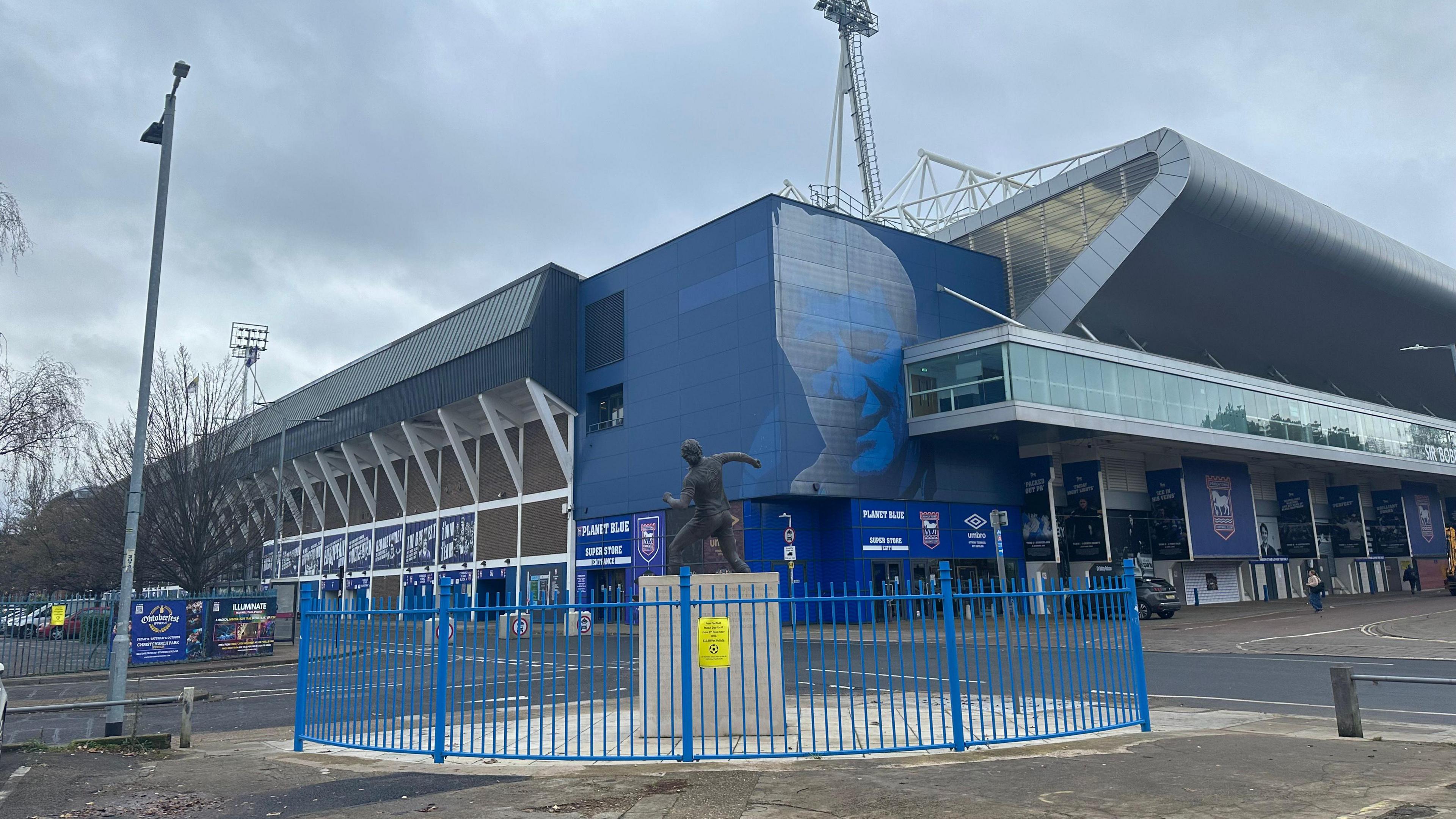 A view of Ipswich Town's home stadium Portman Road. The stadium is largely blue and has branding of the club painted on its exterior. A statue of a player sits in front of the stadium surrounded by a blue fence.