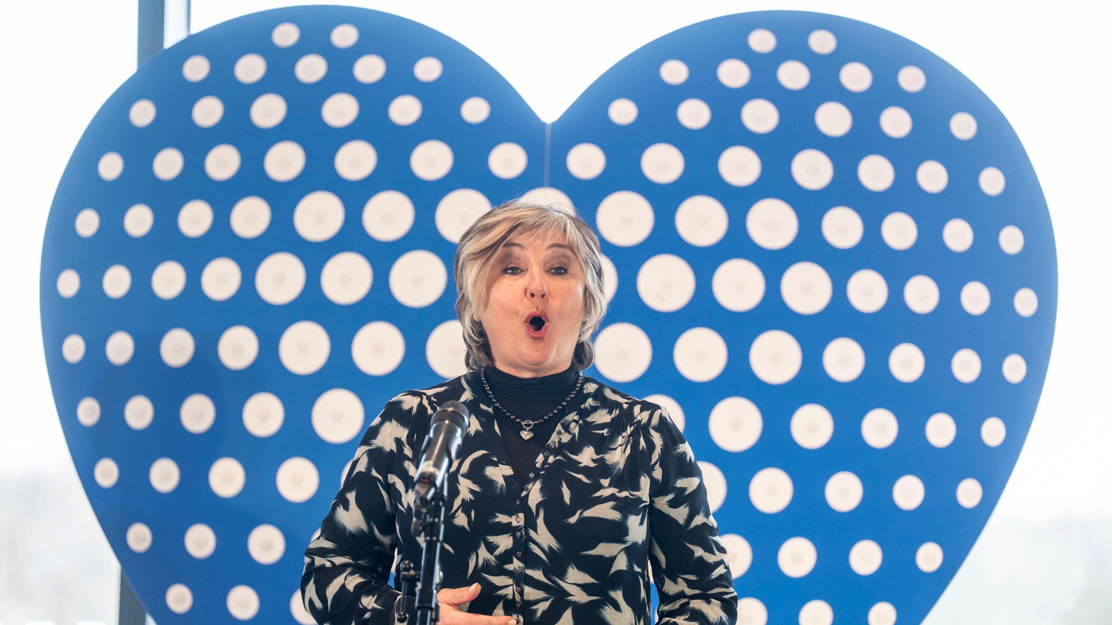 Lesley Garrett, a woman with a black and white patterned jacket, is stood in front of a blue heart shape. She is singing into a microphone in front of her.