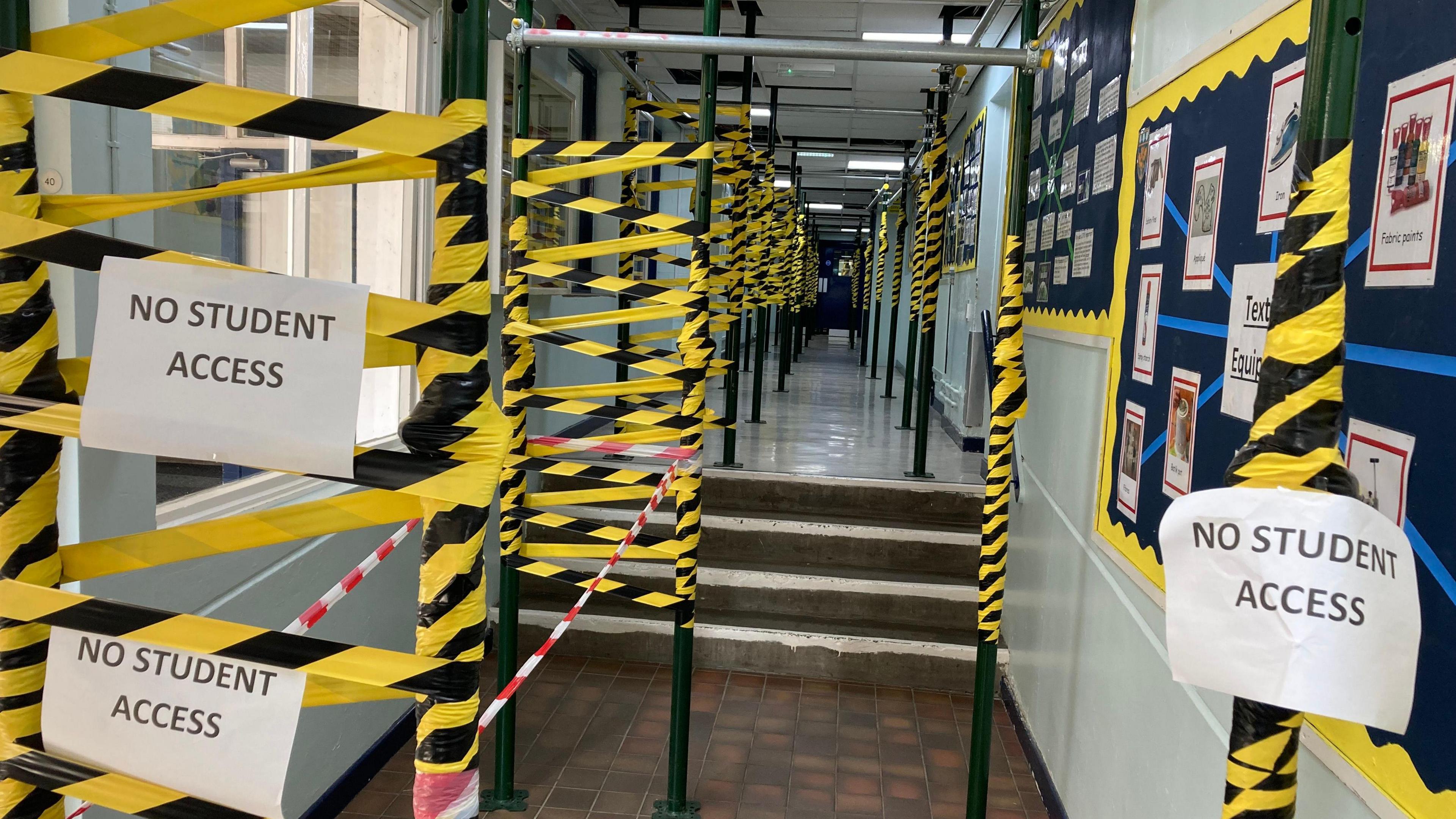 A school corridor, on the left there are black metal poles from floor to ceiling holding up the roof up. They are covered with black and yellow hazard tape and 'no student access signs'. 