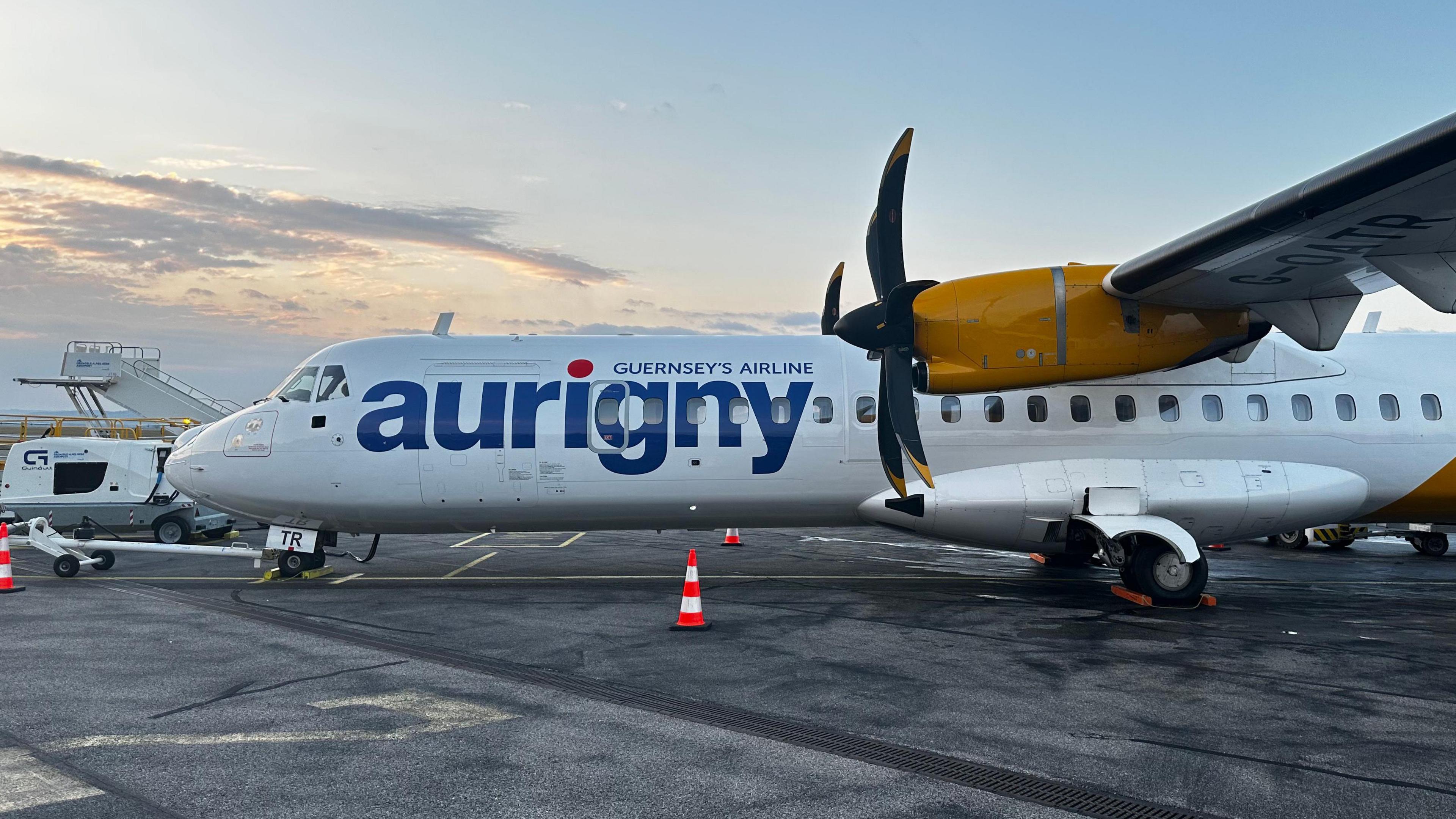 A close up side view of an Aurigny aircraft. The plane is white and yellow. Aurigny is written down the side. 