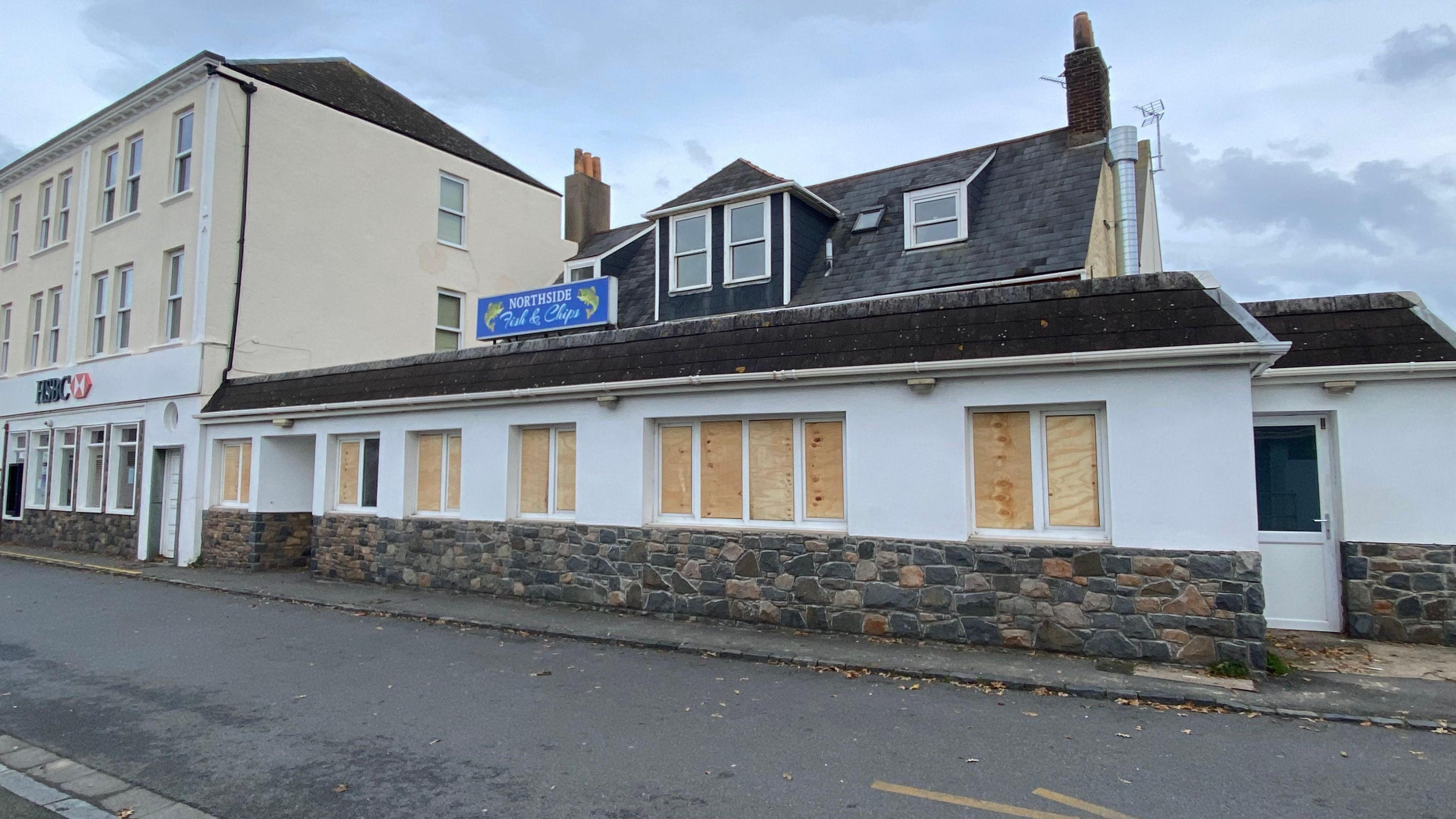 Boarded up windows at the front of Northside Fish and Chips shop. The building is white and has a small blue sign located on top at the centre. There is a brick wall at the front and a door to the right.