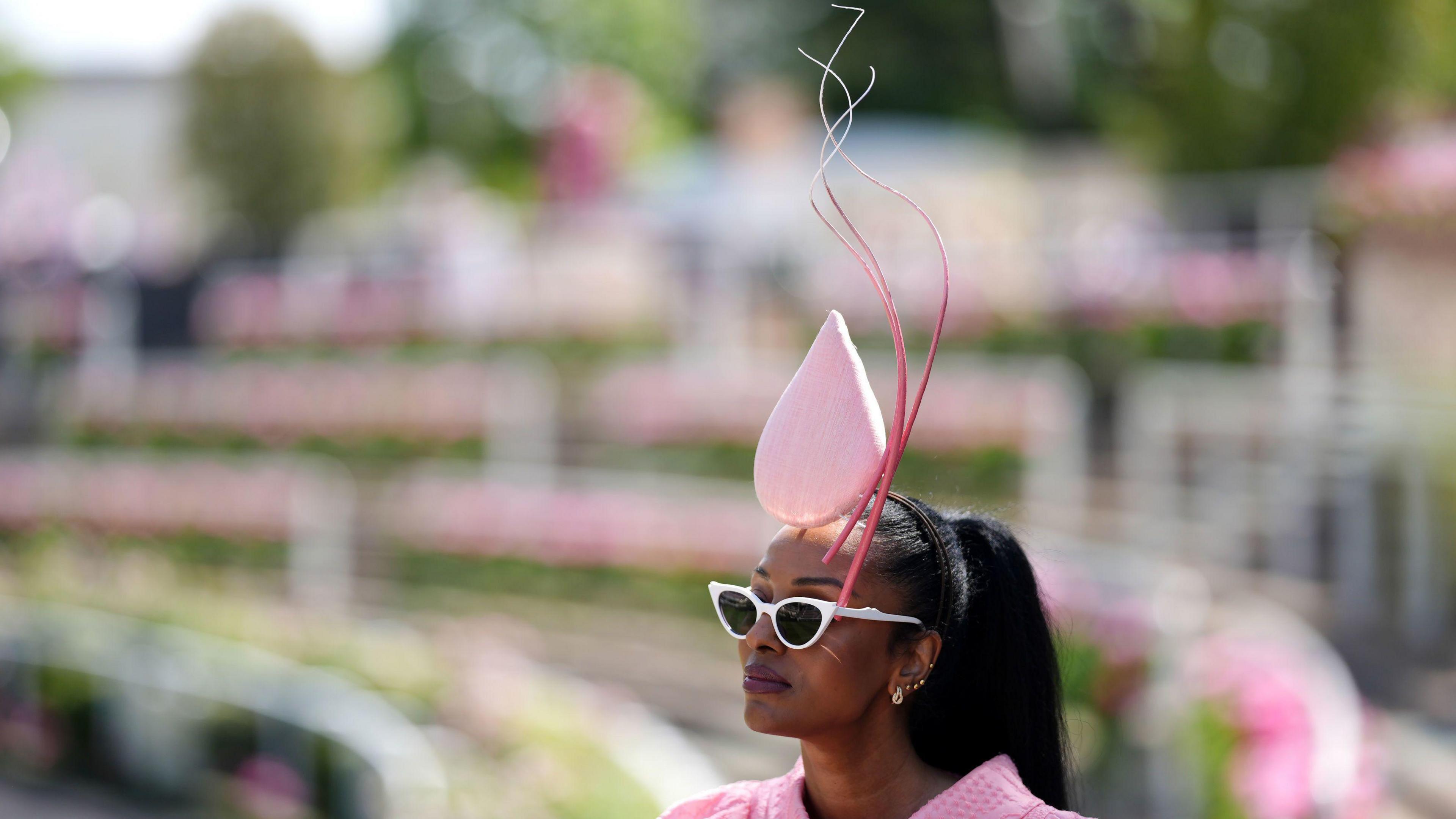 A woman wearing a pink fascinator is pictured in front of pink flowers, which are blurred.
