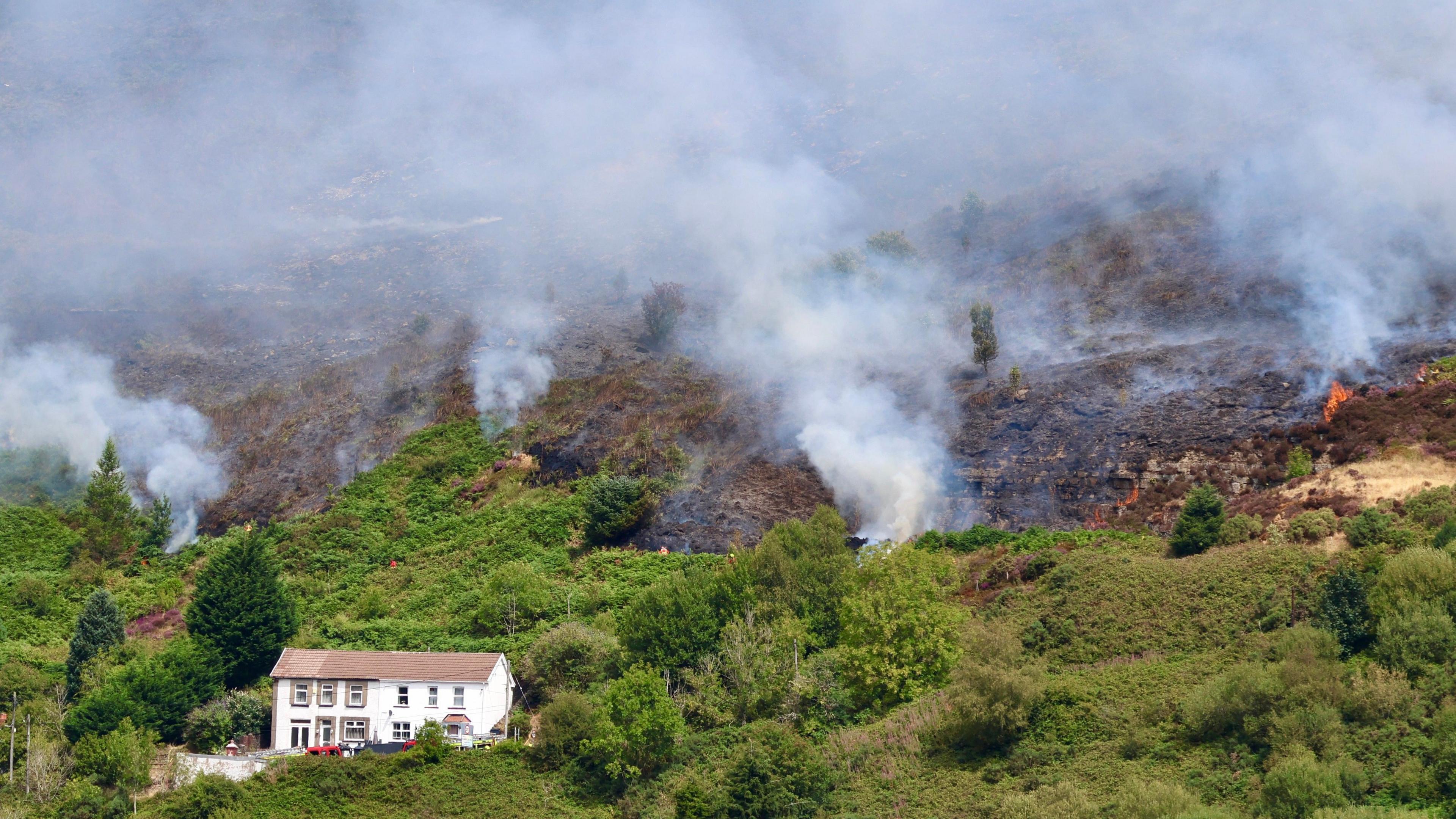 White smoke coming out of fires on a green valley. There is a white house with a terracotta roof in the foreground of the photo