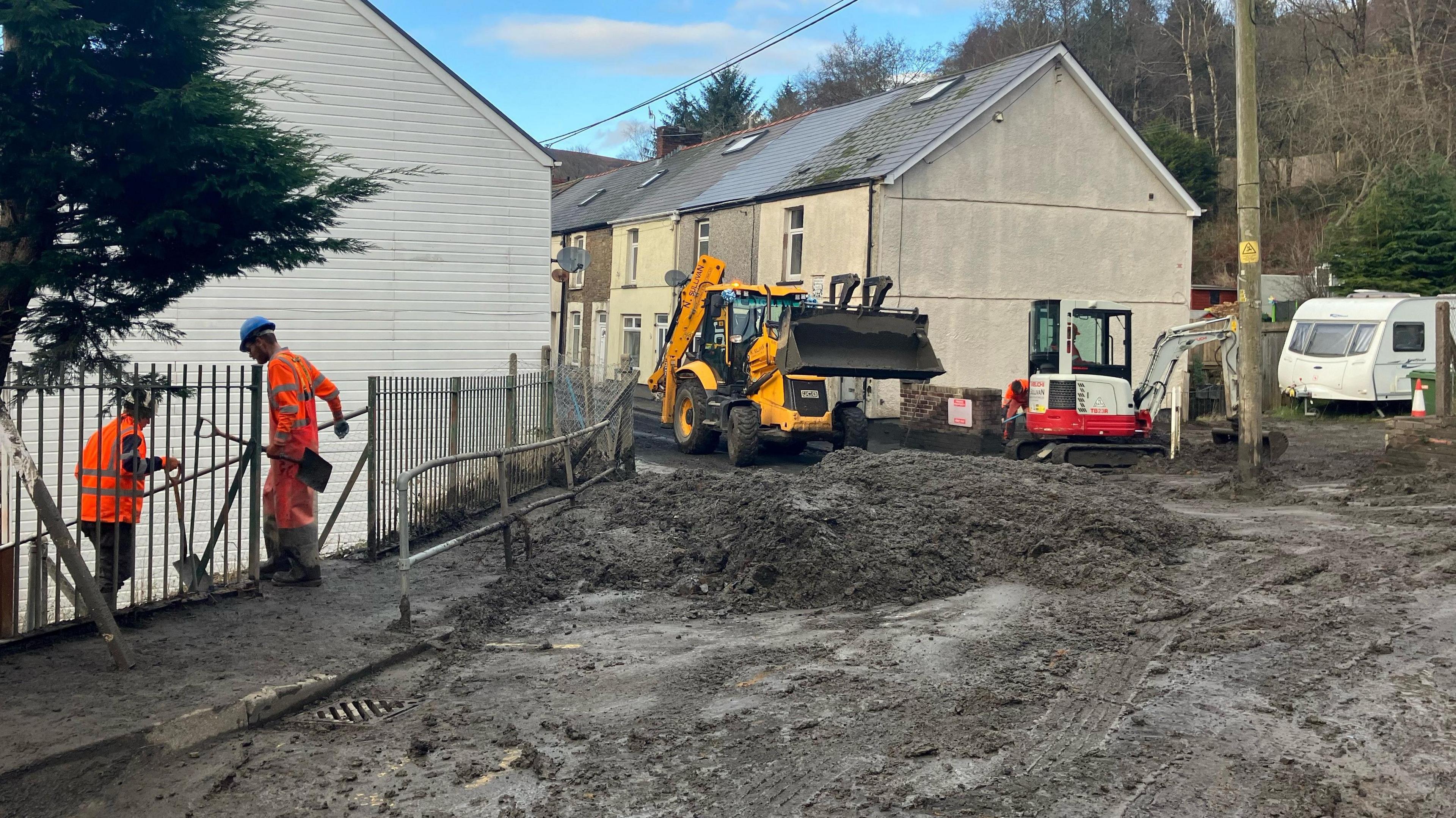 A street in Cwmtillery covered in black slugged piled to make it impassable. Two men in high vis orange jumpsuits and hard hard carry shovels as they stand on the pavement. A yellow digger with a large scoop and a smaller white and red digger with a small scoop are working to move the debris 