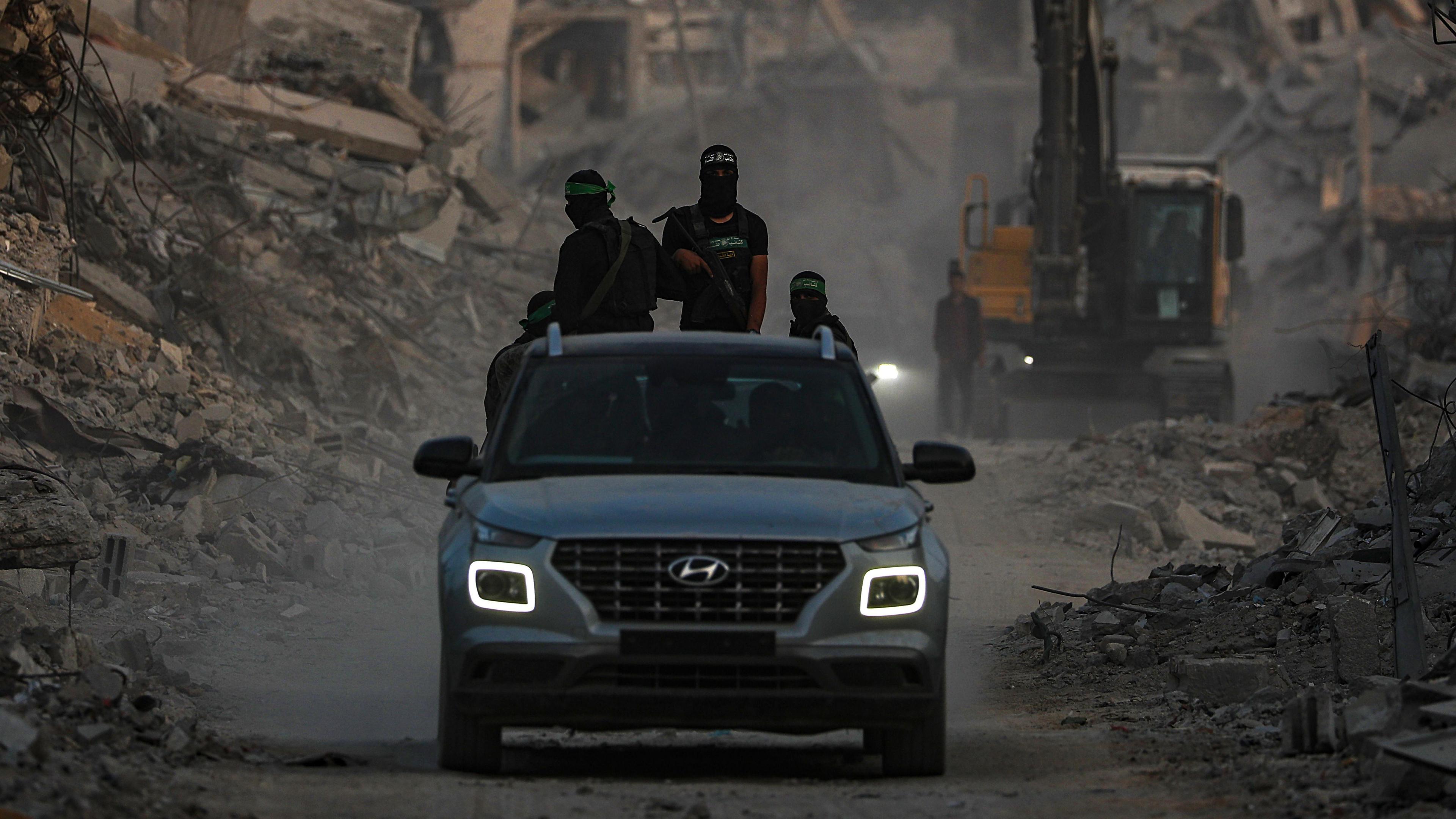 Fighters of the Al-Qassam Brigades, the military wing of the Hamas movement, wearing balaclavas and headbands, and carrying guns, stand guard behind a car as they search for the bodies of Israeli hostages alongside Red Cross workers in Al Shejaeiya neighbourhood in the east of the Gaza City, Gaza Strip. A bulldozer is seen among rubble in the background.