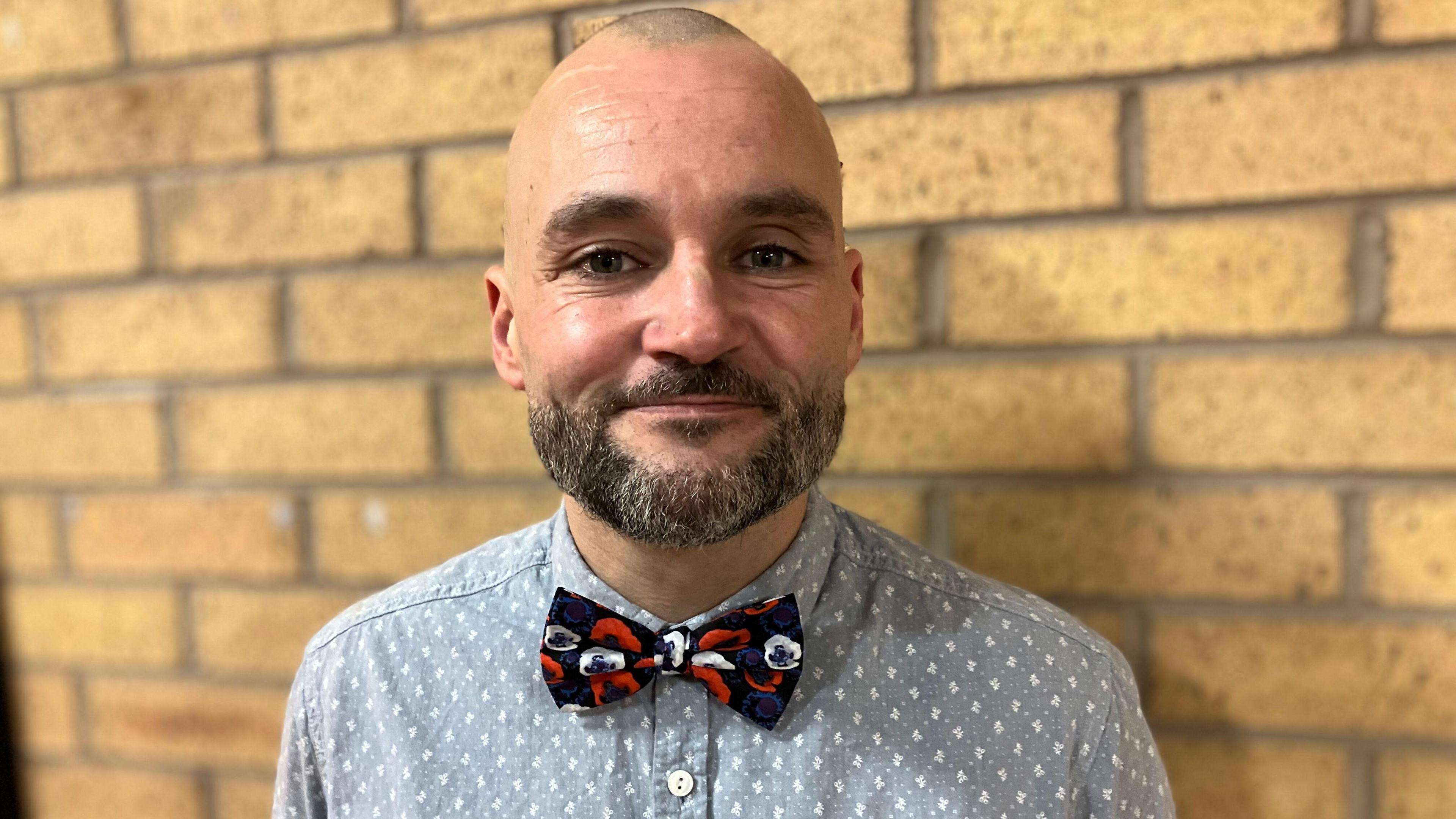 A bald man wearing a grey shirt with a white pattern and a red black and white bow tie, smiles as he stands in front of a brick wall.