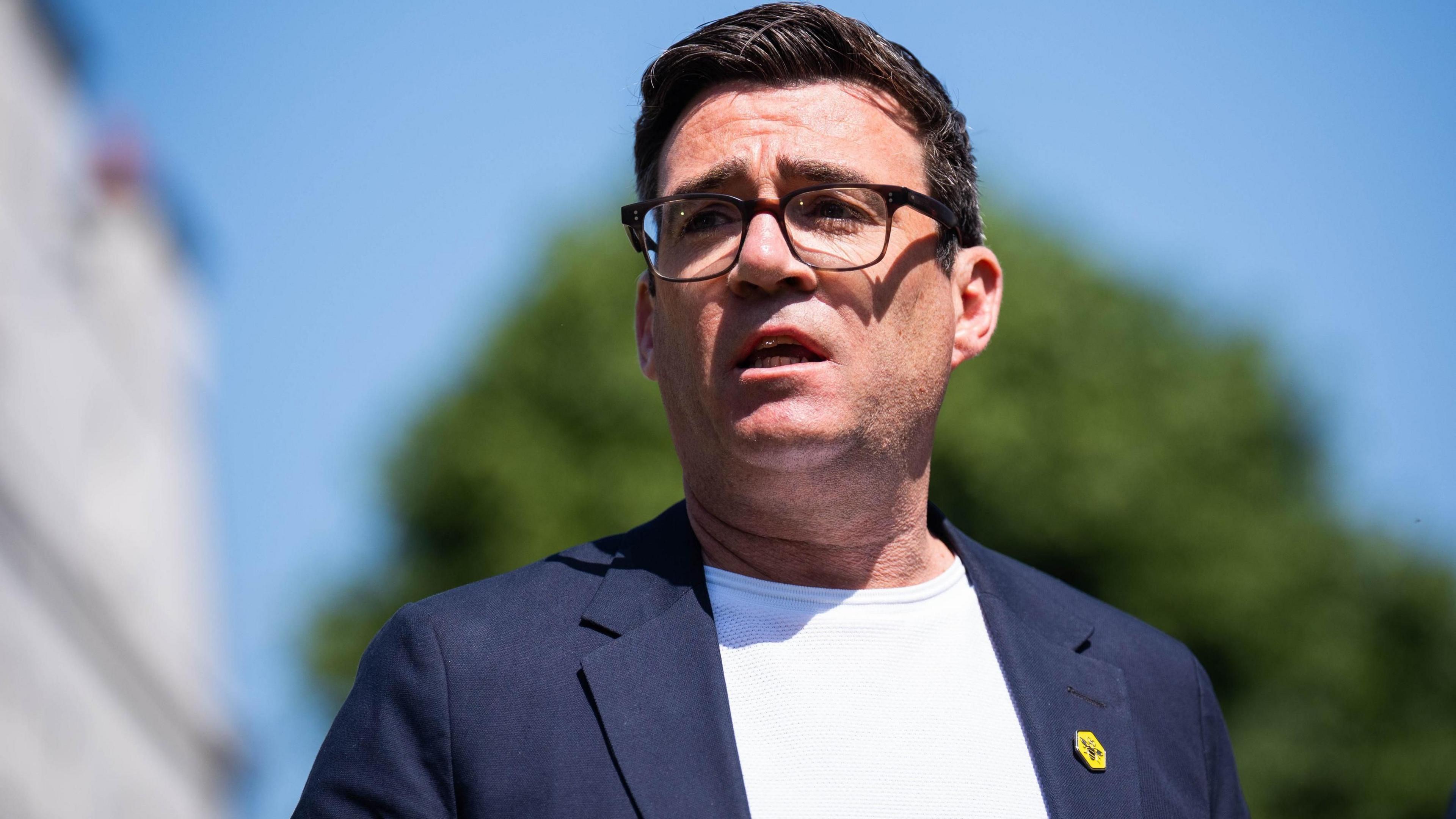 Andy Burnham, who has short dark hair and glasses and is wearing a blue suit jacket with white t-shirt. He has a yellow worker bee badge on his lapel
