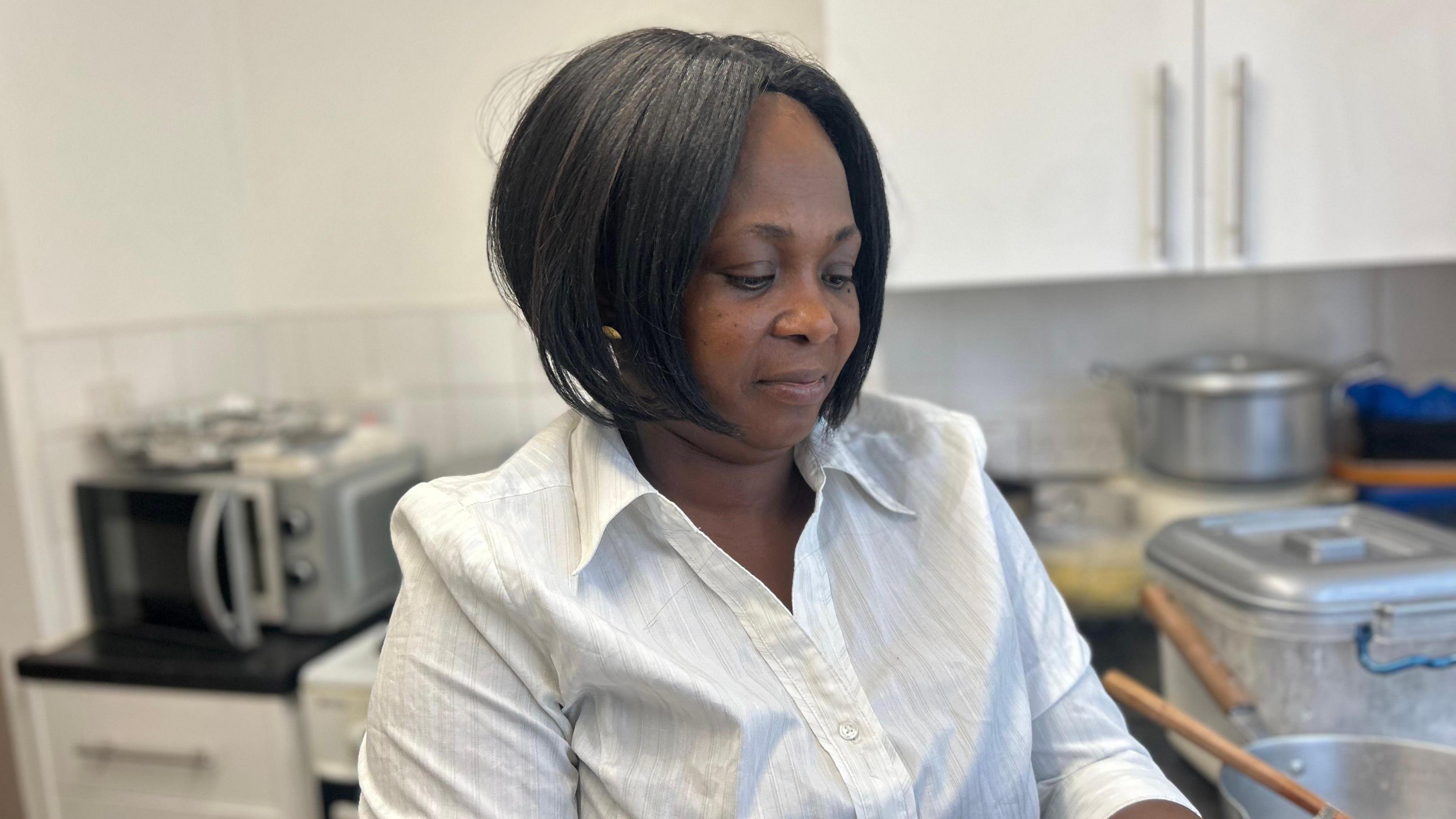 Felicity Afriyie, how has her dark hair cut into a bob and is wearing a white shirt. She sits in a kitchen and is looking down, not at the camera