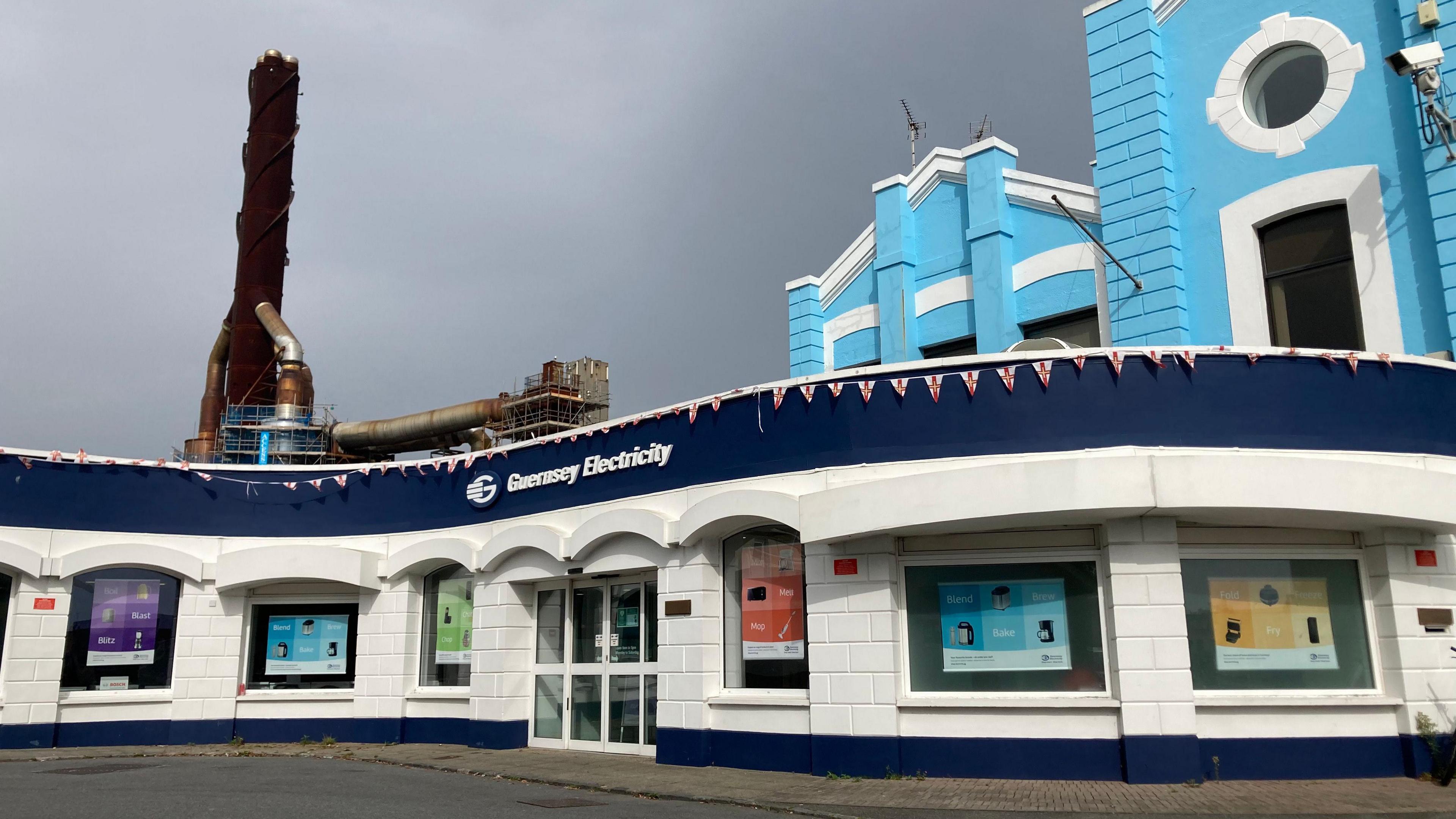 Exterior shot of Guernsey Electricity's headquarters. It is a blue and white building. A large copper chimney is in the background. Grey clouds are in the sky.