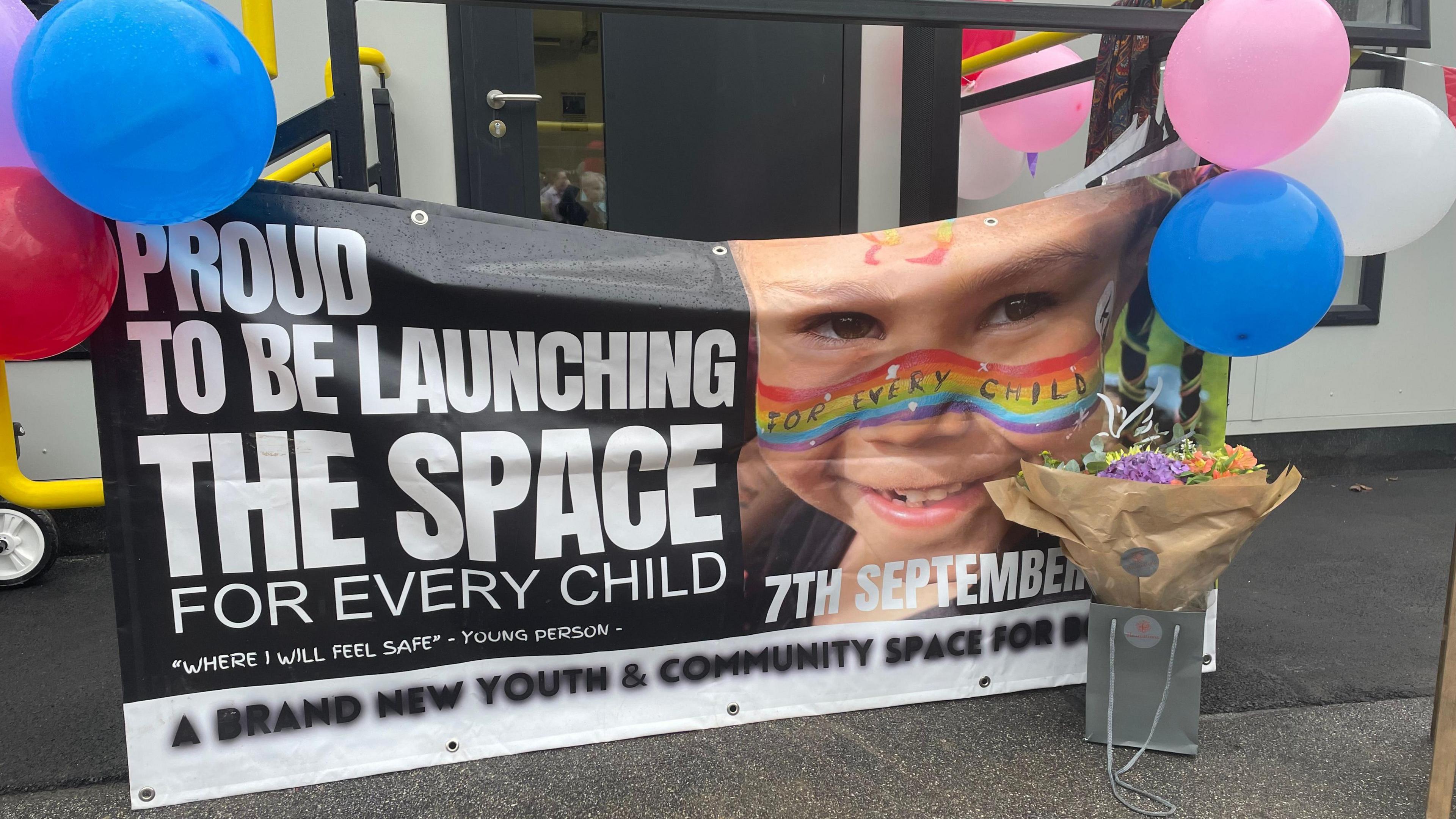 A banner outside The Space with balloons pinned to either edge of the banner and a bouquet of flowers in front.