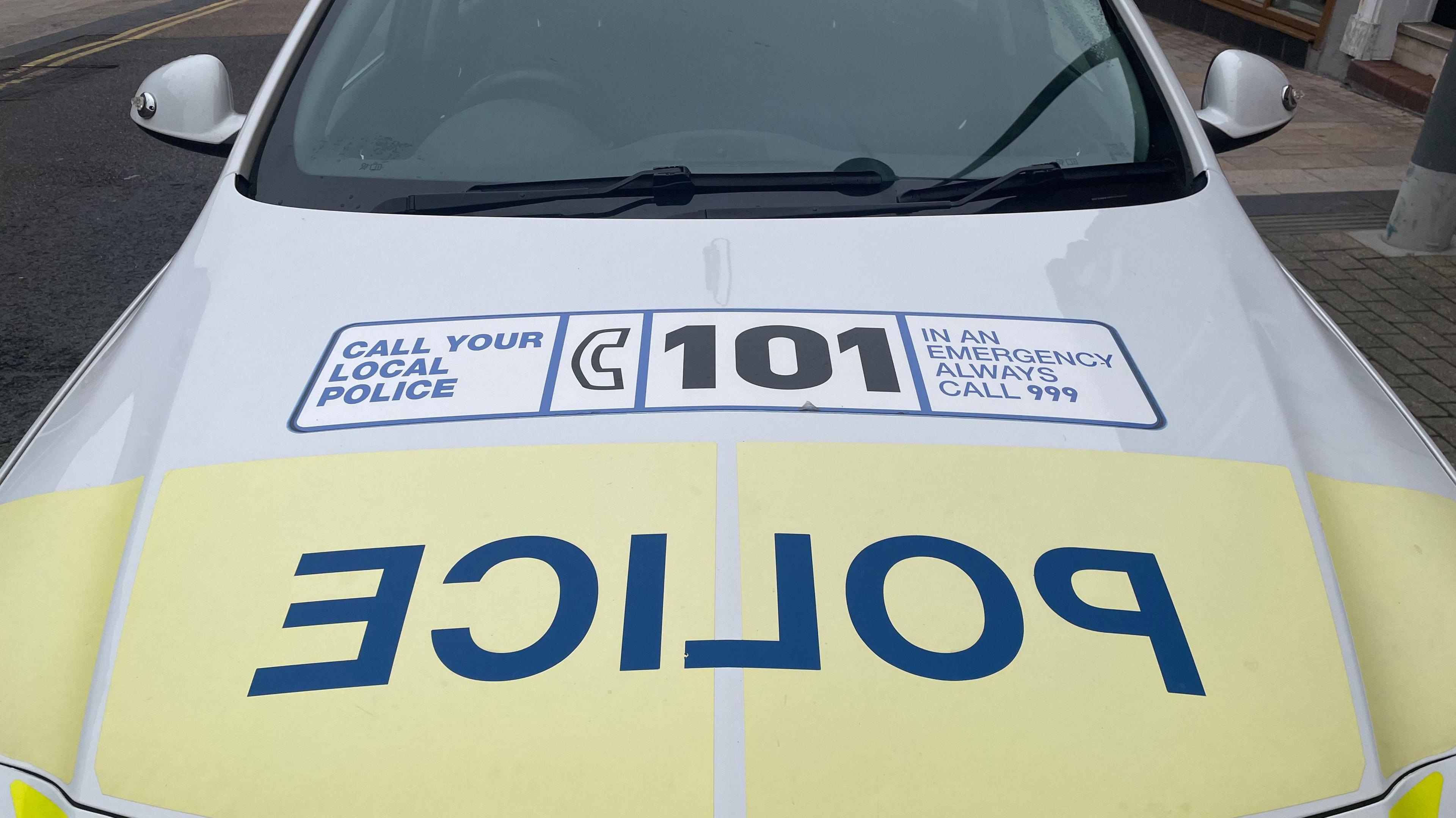 A yellow and white police car with police written in large blue letters on the front in reverse and a sticker on it saying call 101.