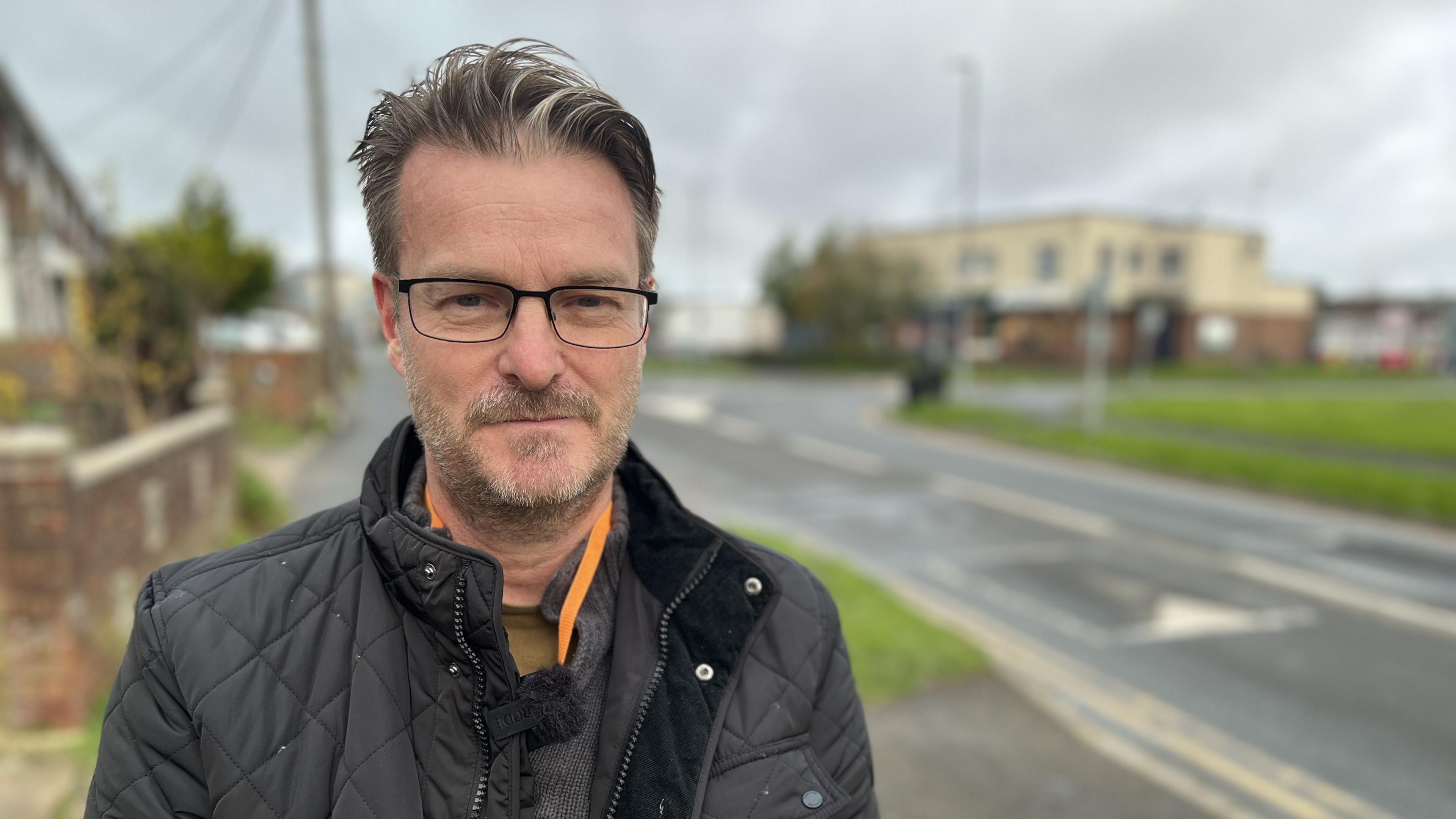 Swindon Borough Council member Chris Watts standing next to a road wearing black rimmed glasses and short grey hair and beard. He is wearing a padded black jacket