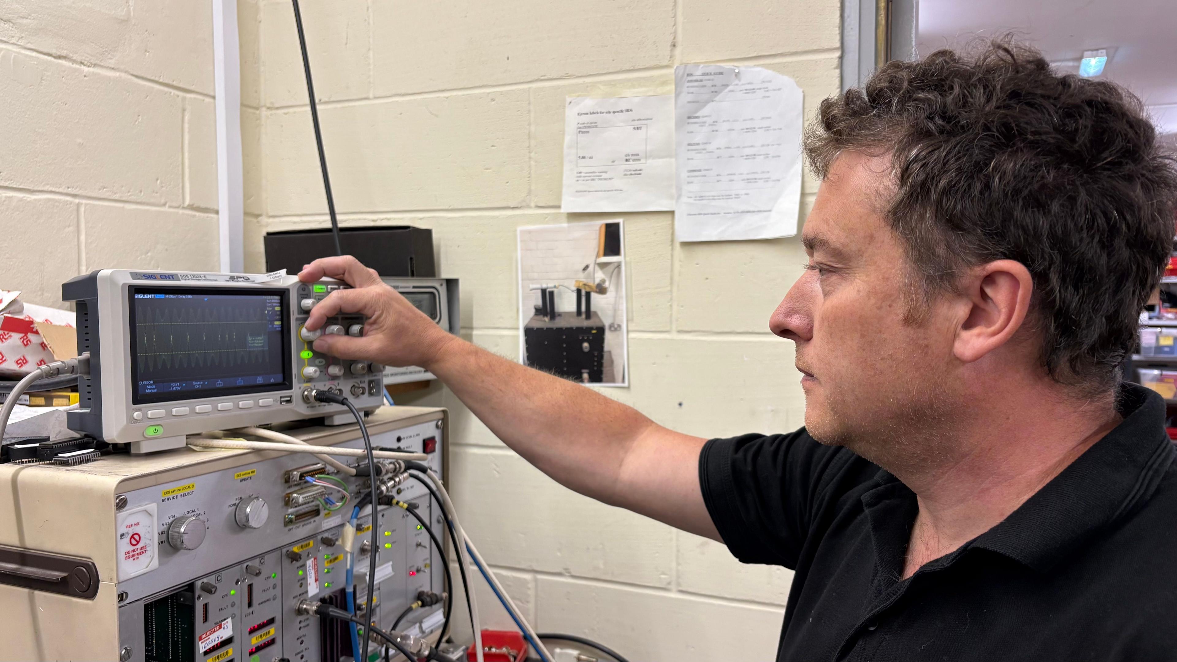 Phil Critchley with short dark curly hair, wearing a black T-shirt. He has one arm stretched towards a small piece of equipment with a screen on the front. He is adjusting a setting on the equipment. There is more equipment beneath and to the right. There is a light-coloured brick wall behind him.