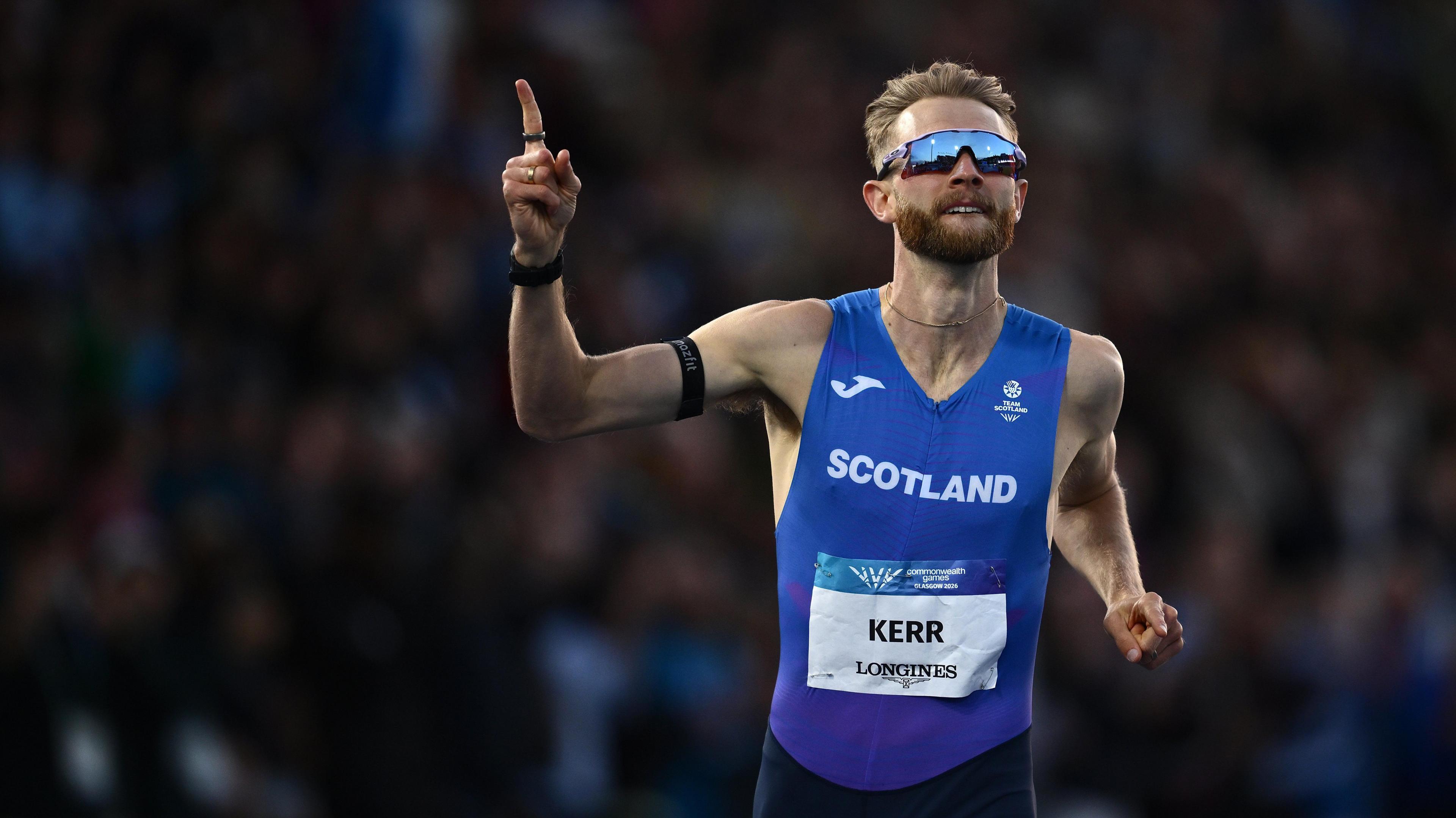 Josh Kerr wearing sunglasses and a blue running vest, holding a finger to the sky