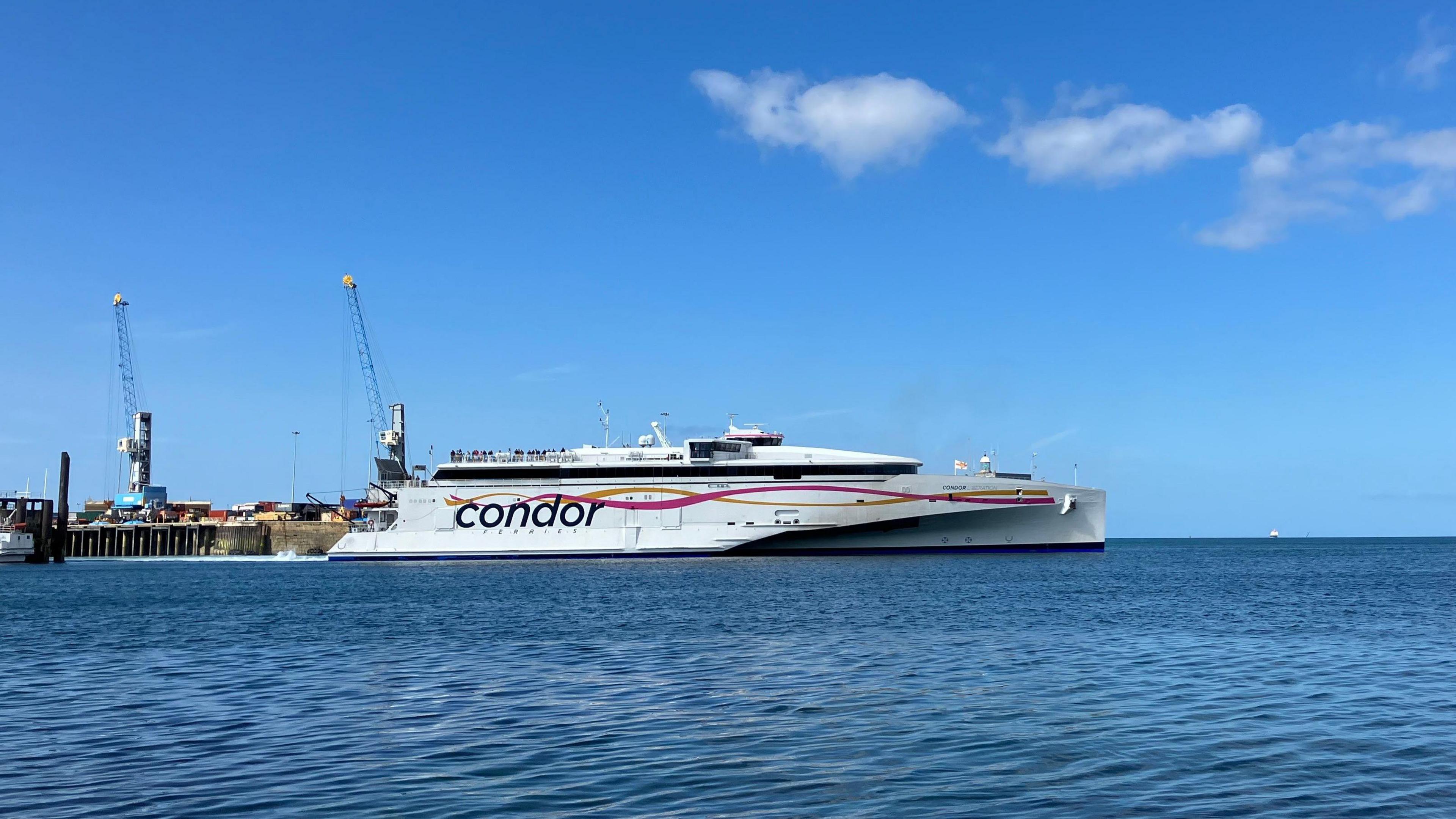 A large, white trimaran ferry with the word Condor emblazoned on the site, along with yellow and pink ribbons, pulls away from an old granite pier on a sunny day.