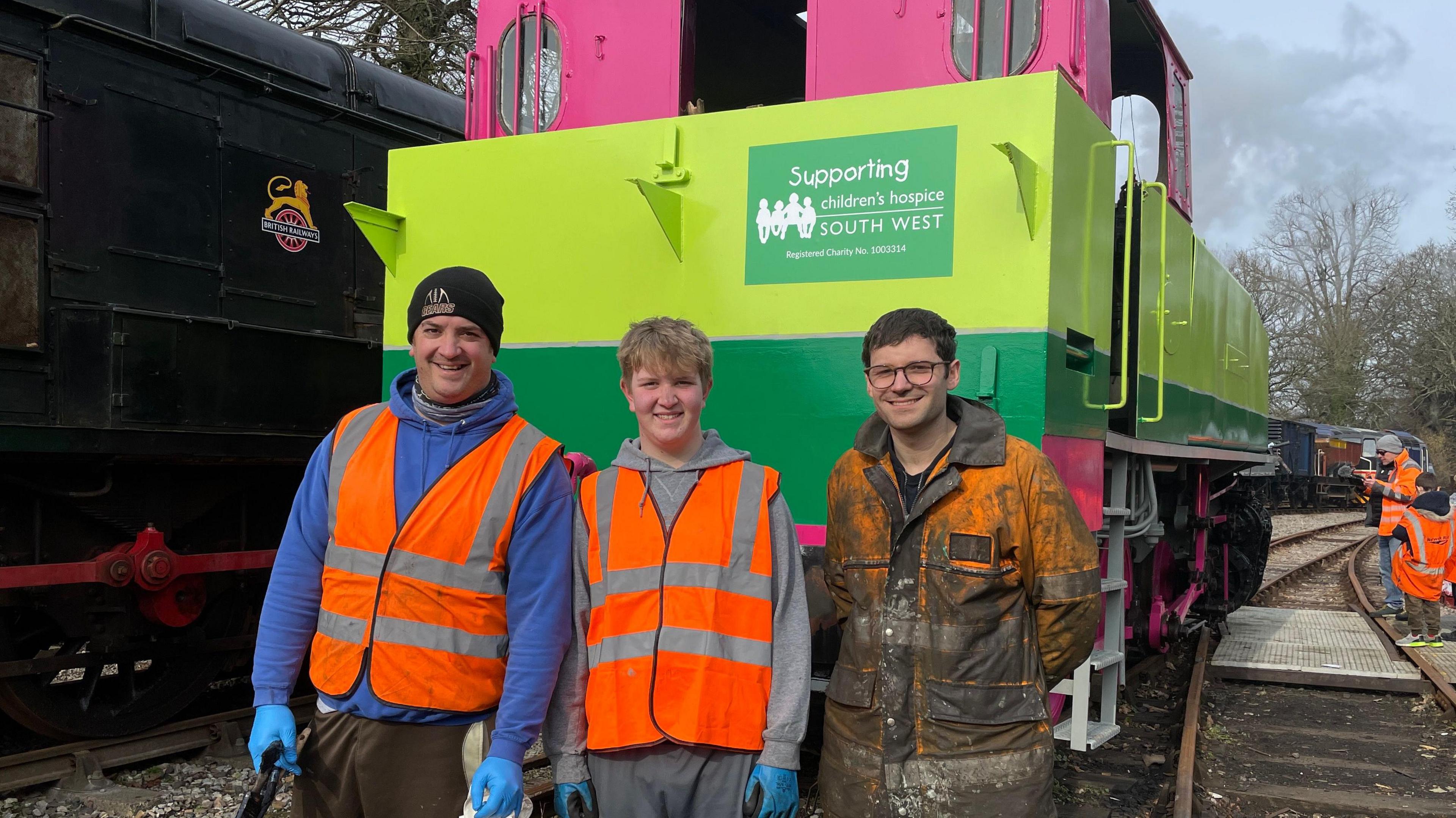 Three of the people in the restoration team, Oliver's dad, Mike Brown, his brother Ben, and Marc Bellin in orange overalls, stand in front of the bright green and pink train. 