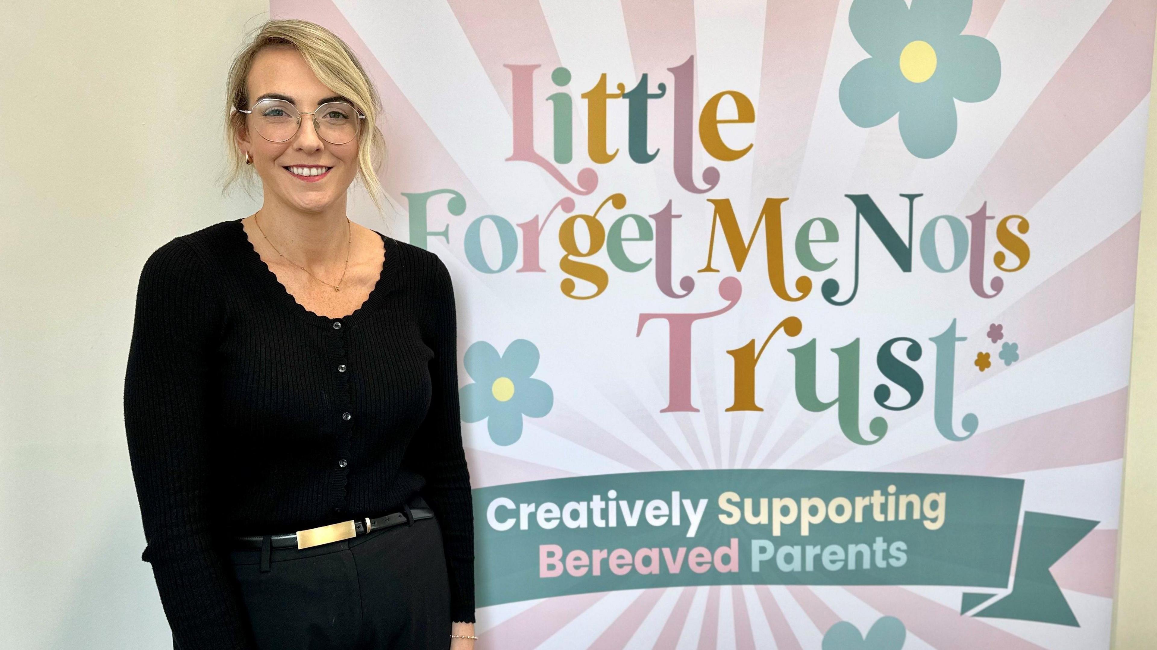 A blonde haired woman with glasses stands in front of a billboard with Little Forget Me Nots Trust on it. 
