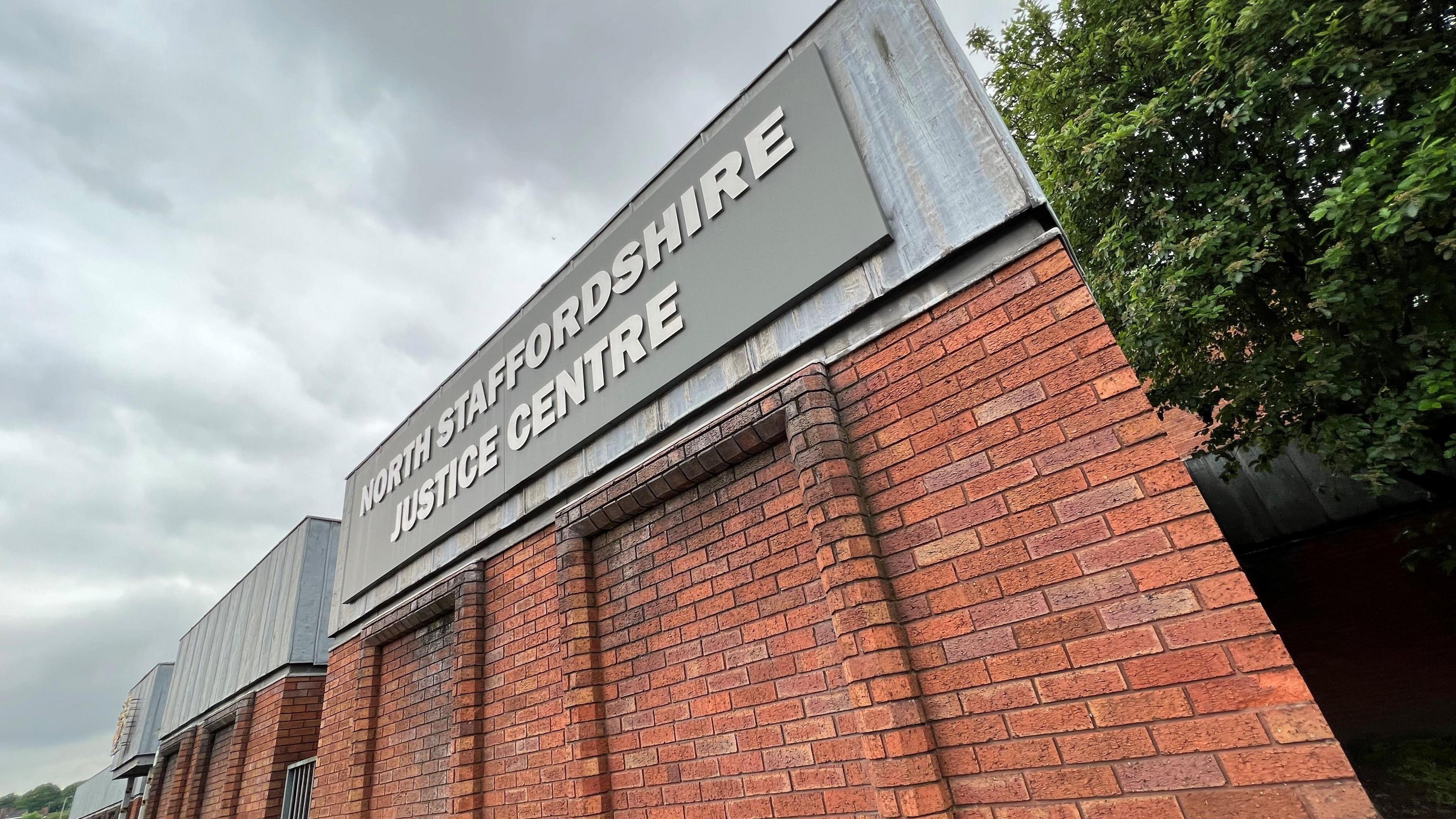 A red brick building, viewed from a corner, with metal flashing around the top. There is a grey sign with lettering that reads North Staffordshire Justice Centre.