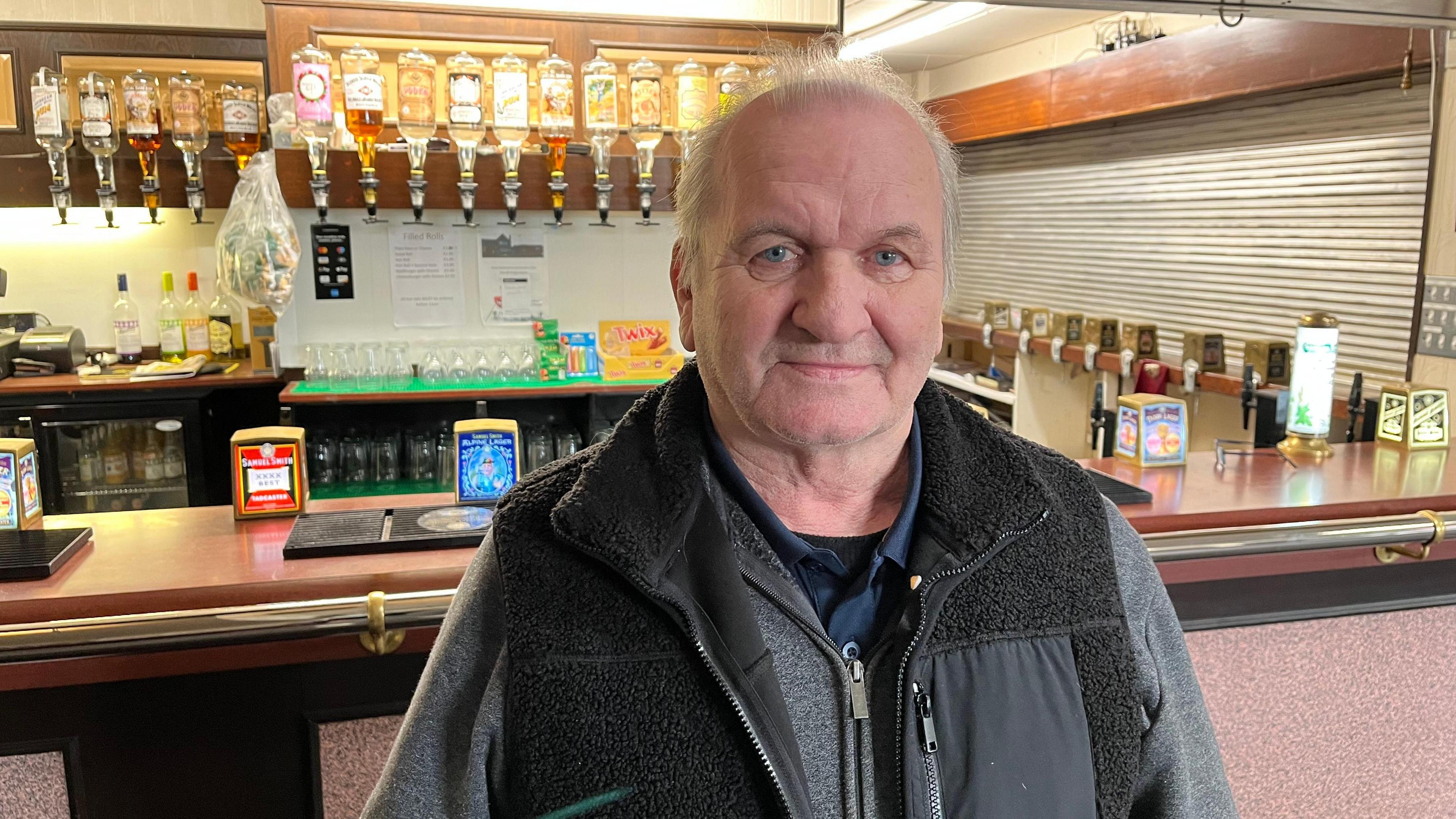 A man in a black coat stands in front of a bar. Behind him are lots of glasses and bottles of alcohol.