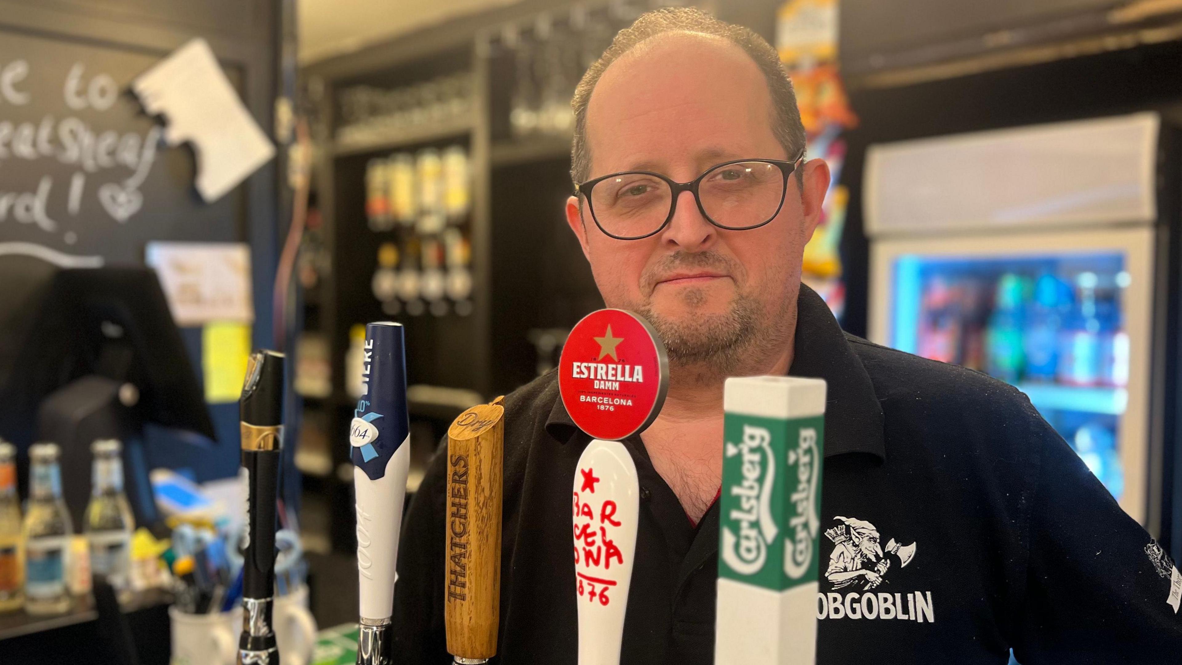 David Sutton stands behind beer pumps in his pub and wears a pair of black-rimmed glasses and a black polo shirt.