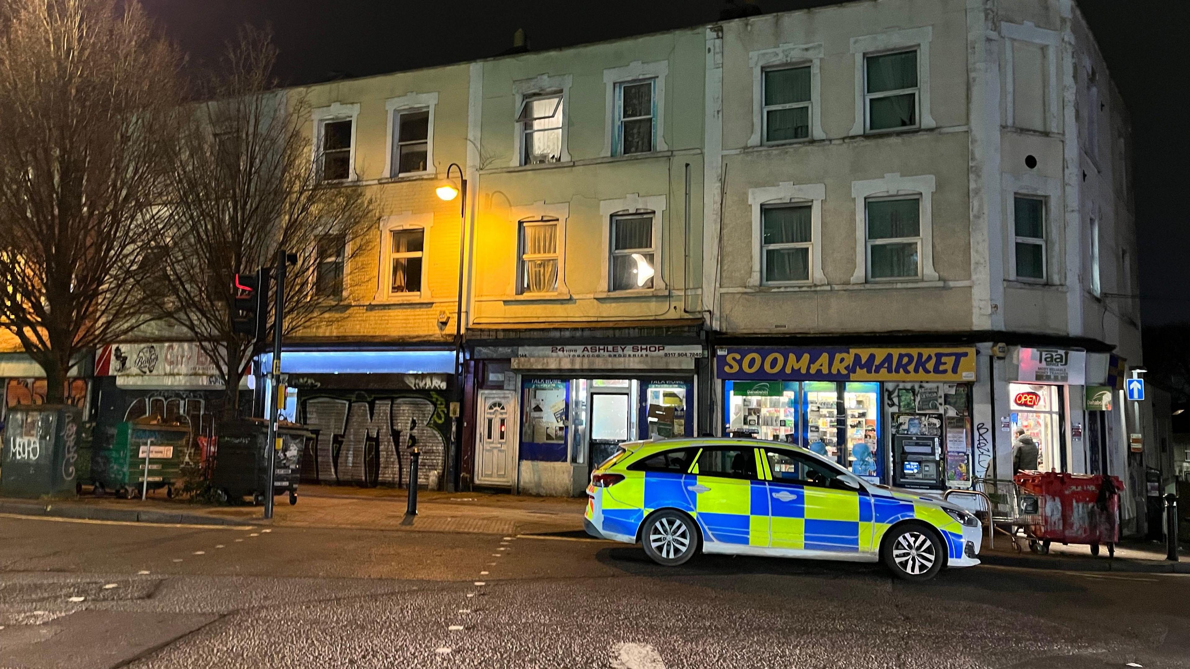 A police car, pictured to the bottom right of the image, parked on a street after dark with the road lit by a lamp-post. Shop fronts can be seen on the ground floor of a row of buildings with windows on the floors above and trees to the left of the image.