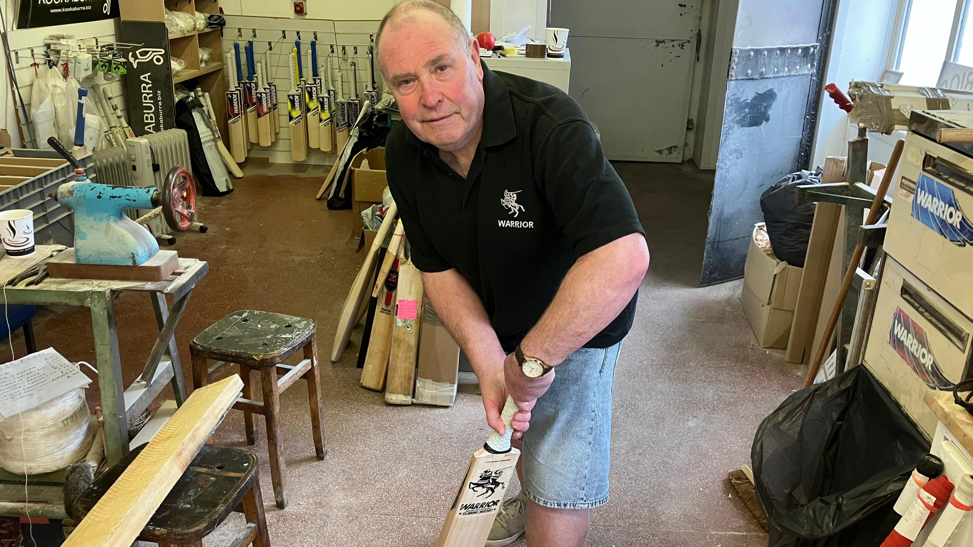Ian Sampson, a man in his 60s, feigns a forward defensive stroke with a cricket bat in a workshop surrounded with cricket bats, tools and benches. He is wearing a black t-shirt and denim shorts.