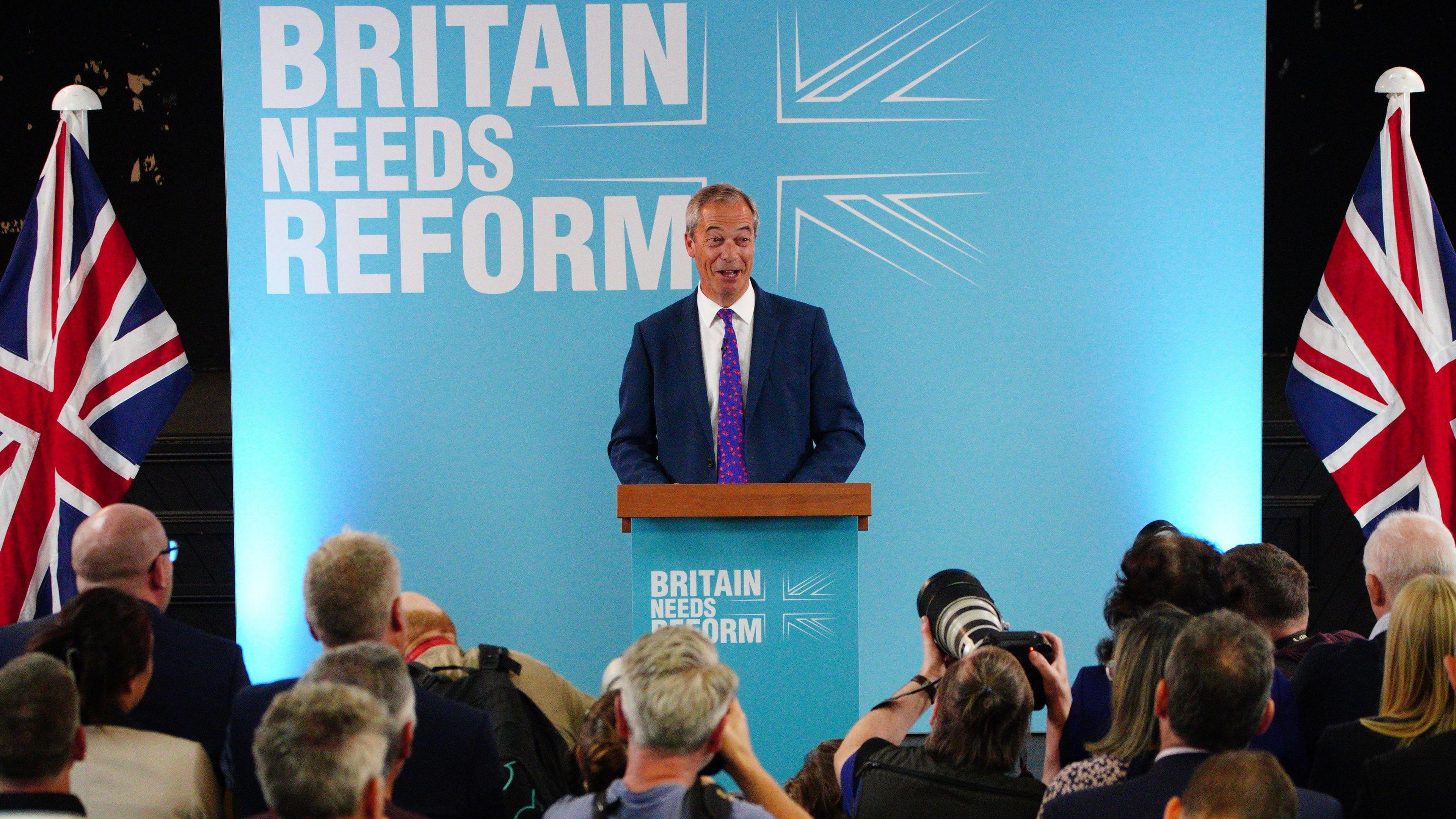 Llun o Nigel Farage ar lwyfan. Mae'n gwenu o flaen rhes o bobl yn pwyntio camera tuag ato. Mae baneri Jac yr Undeb ar naill ochr i'r llwyfan. Mae'r geiriau 'Britain needs Reform' ar boster mawr glas y tu ôl iddo.
