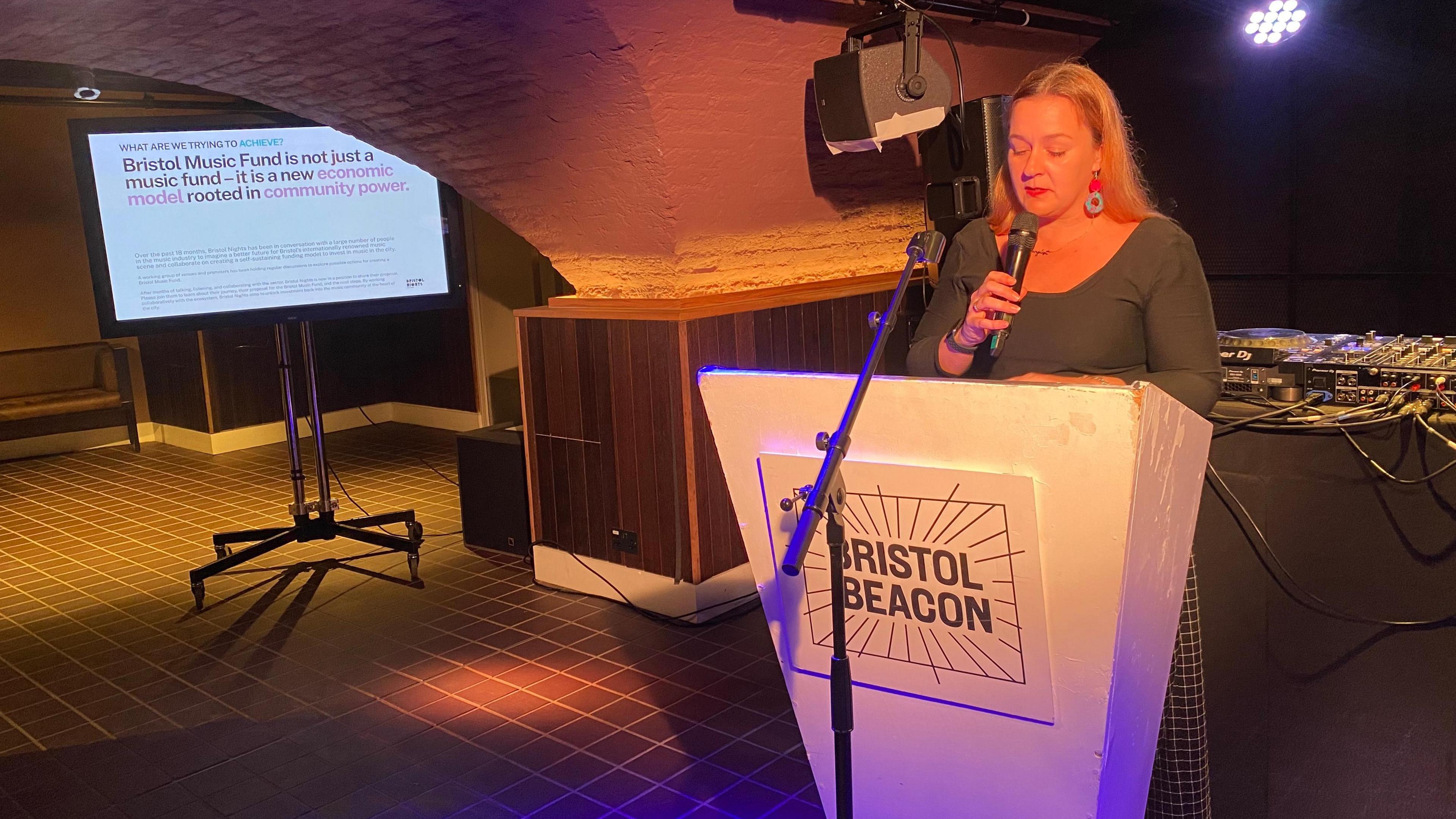 Carly Heath holding a microphone and standing at a white lectern that says Bristol Beacon on it. There is a big screen in the background giving info about the Bristol Music Fund