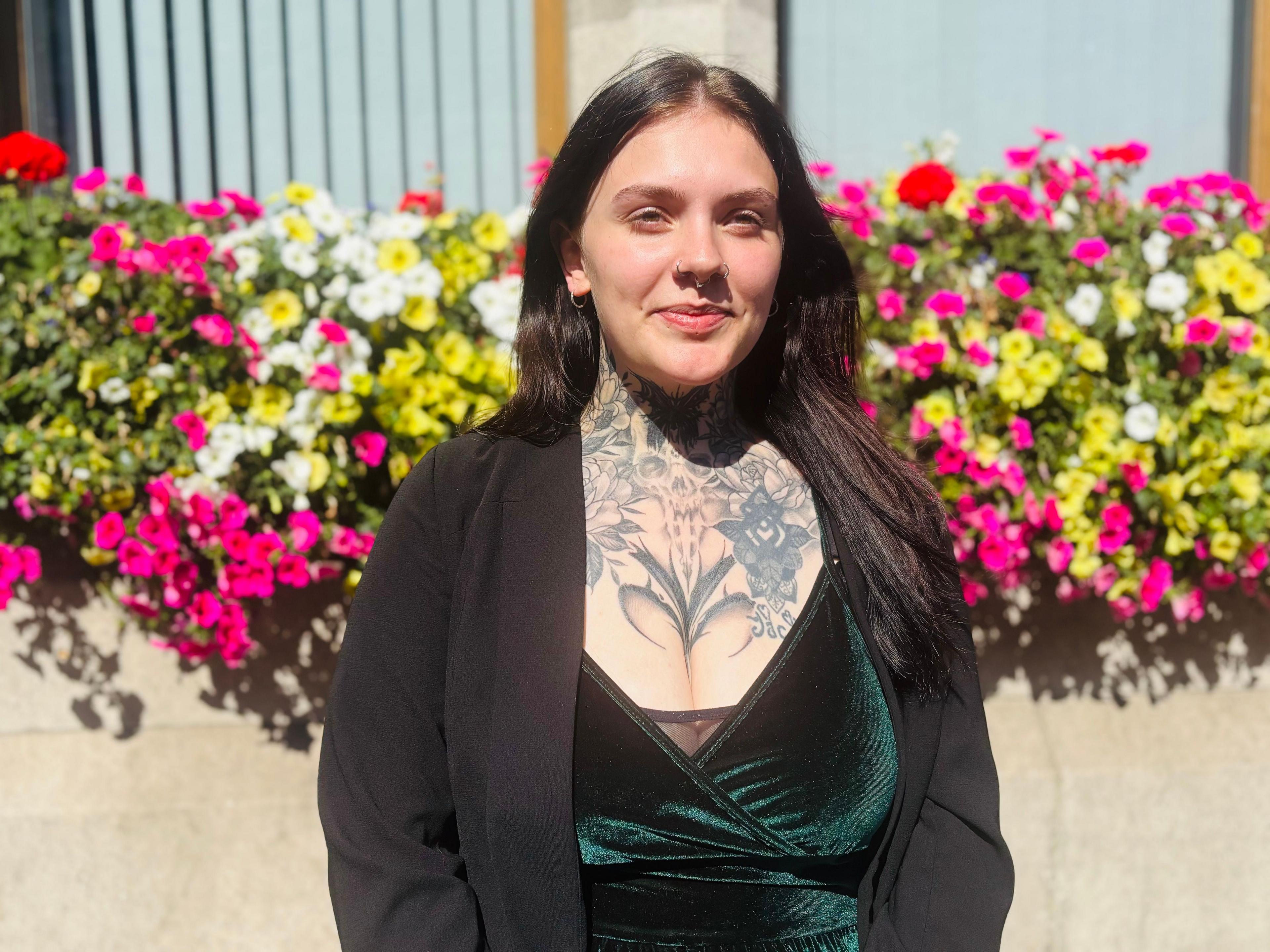 A young woman with dark hair smiling at the camera. She has a large tattoo on her chest and is wearing black clothes. She is standing in front of colourful flowers. 