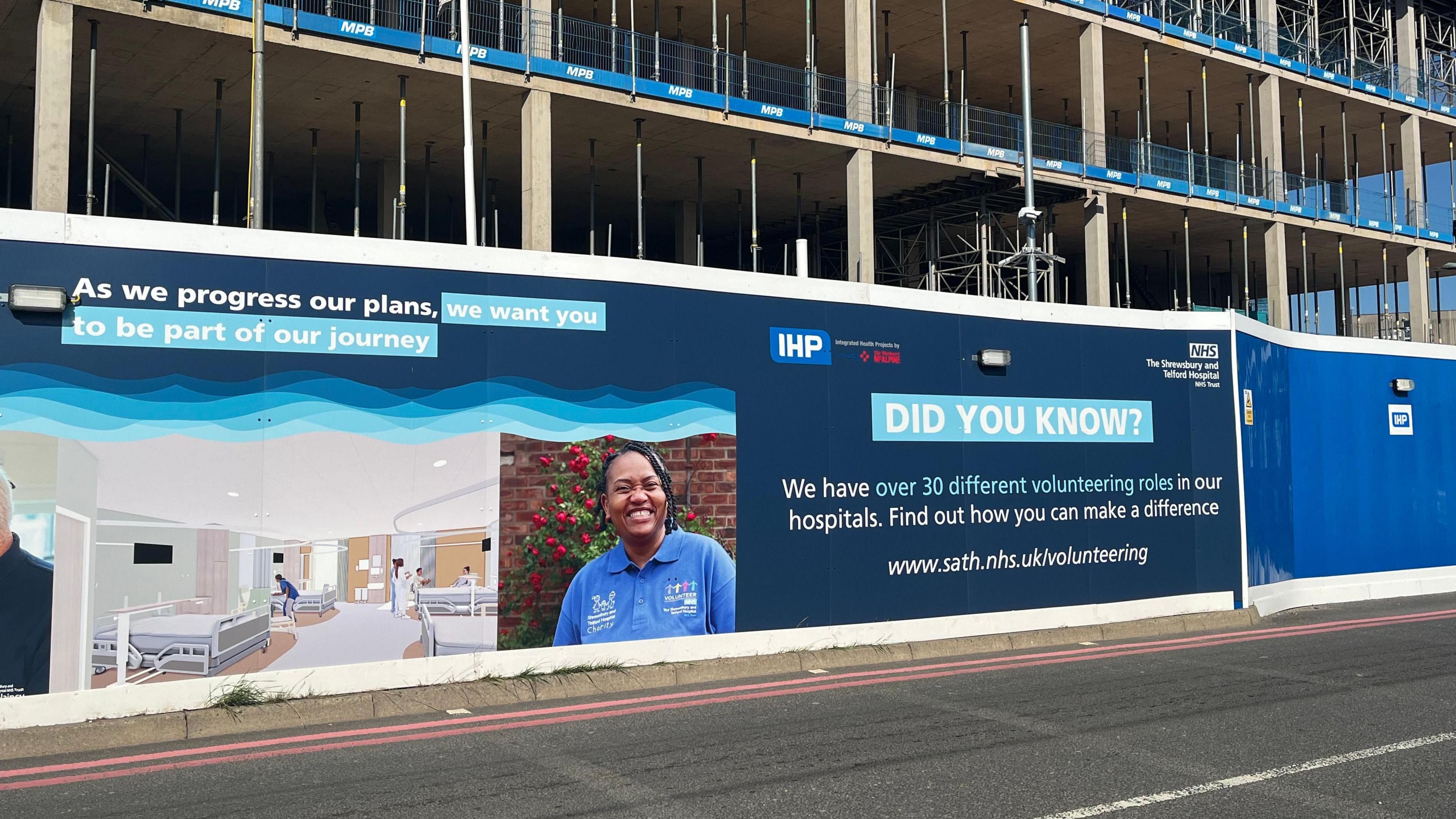 Photo of hoarding for a building site at the Royal Shrewsbury Hospital. It's dark blue, with images of a hospital ward and a staff member to the left. On the right of the billboard, white writingreads: 'As we progress our plans, we want you to be part of our journey. Did you know? We have over 30 different volunteering roles in our hospitals. Find out how you can make a difference: www.sath.nhs.uk/volunteering.'