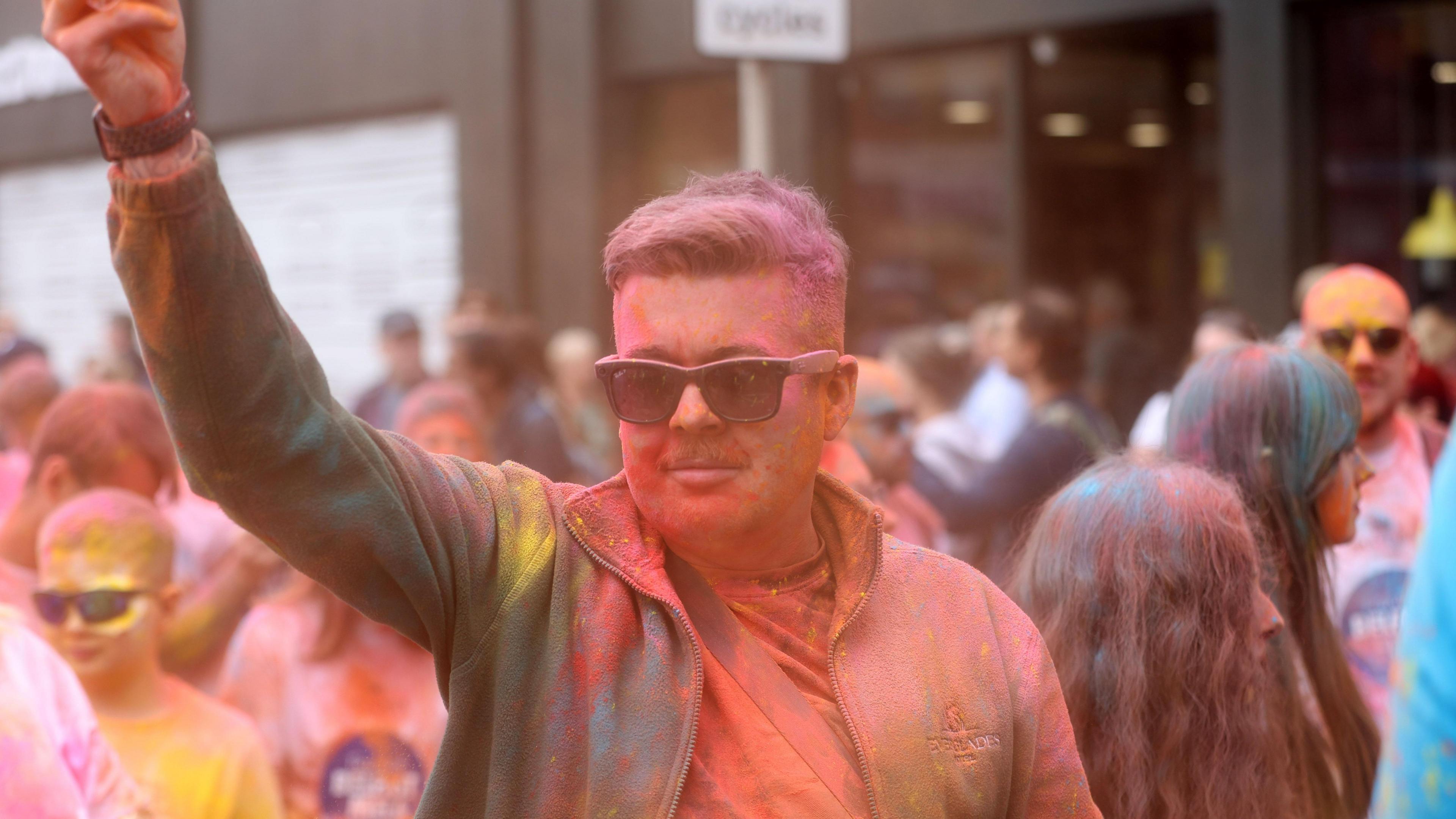 A man is covered in coloured paint powder. He is wearing black sunglasses and has his arm raised in the air.