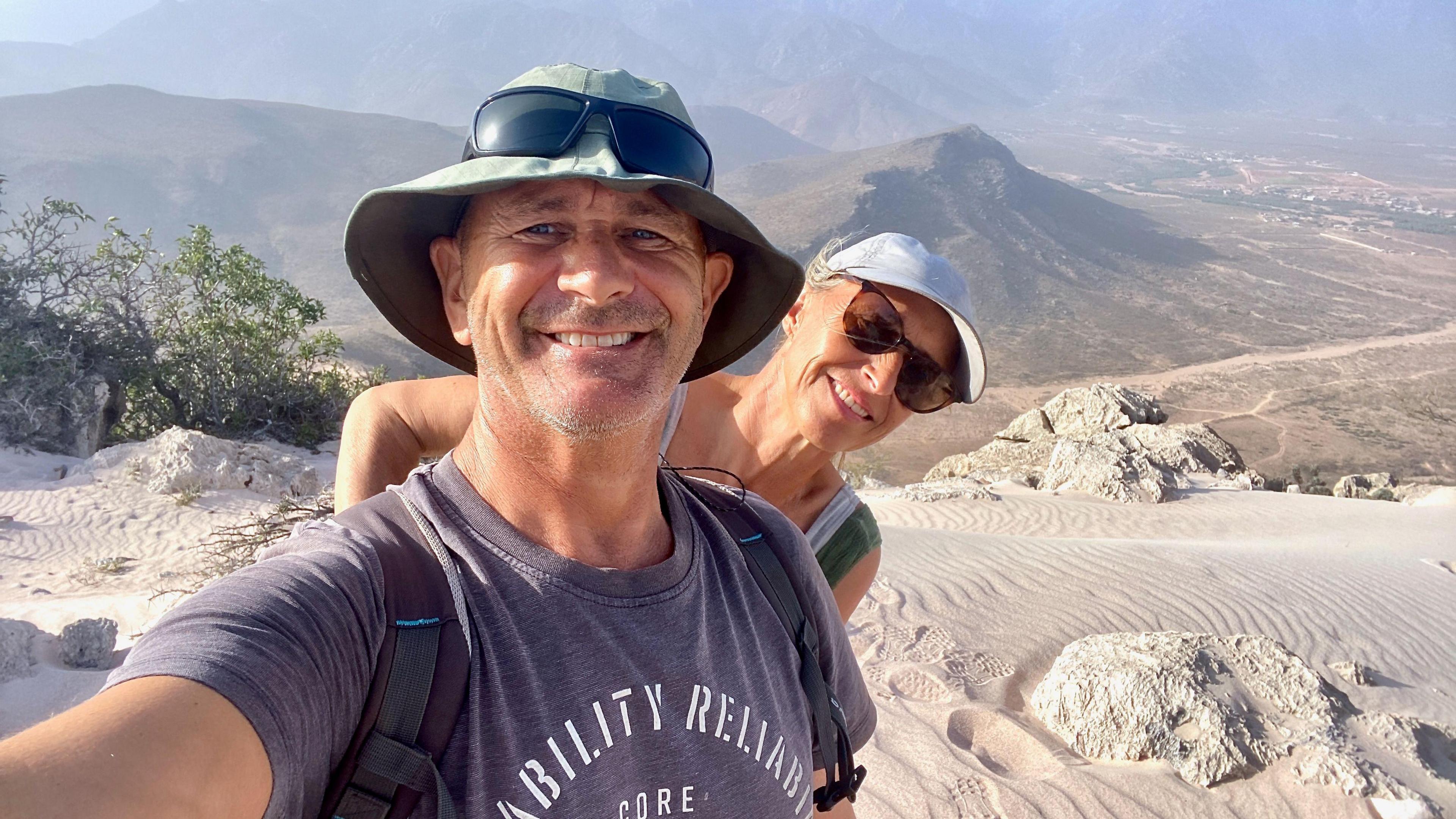 A man wearing a bucket hat and grey shirt. A woman with a cap and sunglasses is standing behind him. They are standing on the desert.