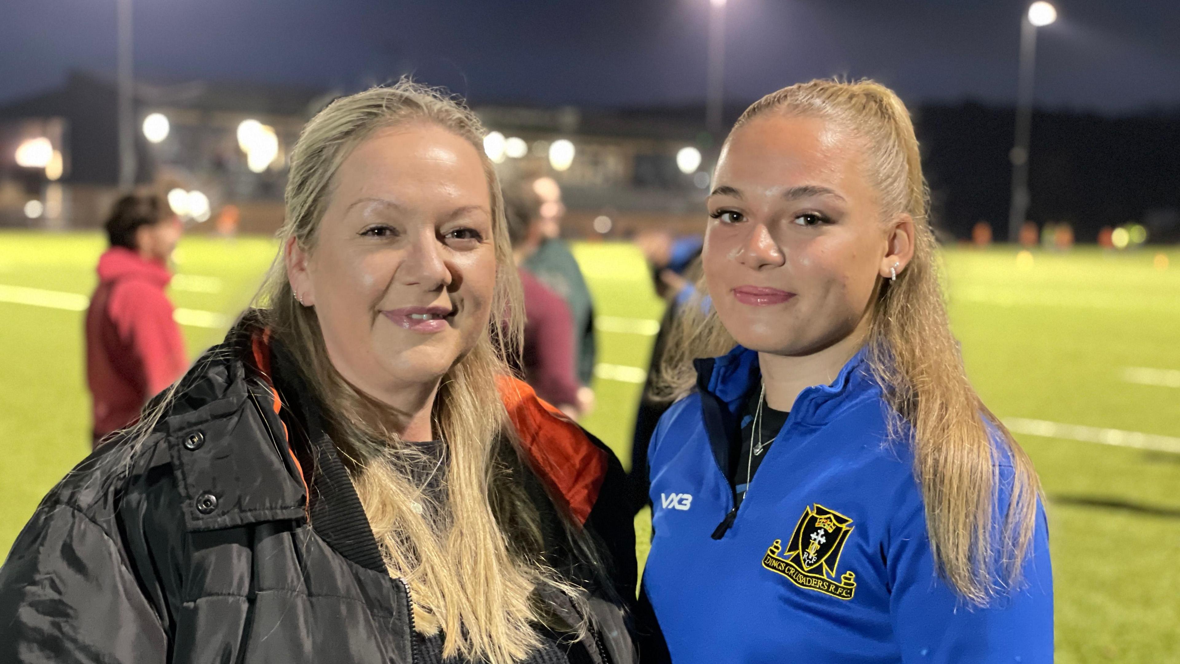 Mother Sam MacDonald (L) stands next to Roxy (R) and they're smiling at the camera. Sam is wearing a black puffer coat and  Roxy wearing a light blue jacket with the Dings Crusaders RFC logo on the right side. Behind them is a blurred image of a rugby pitch.