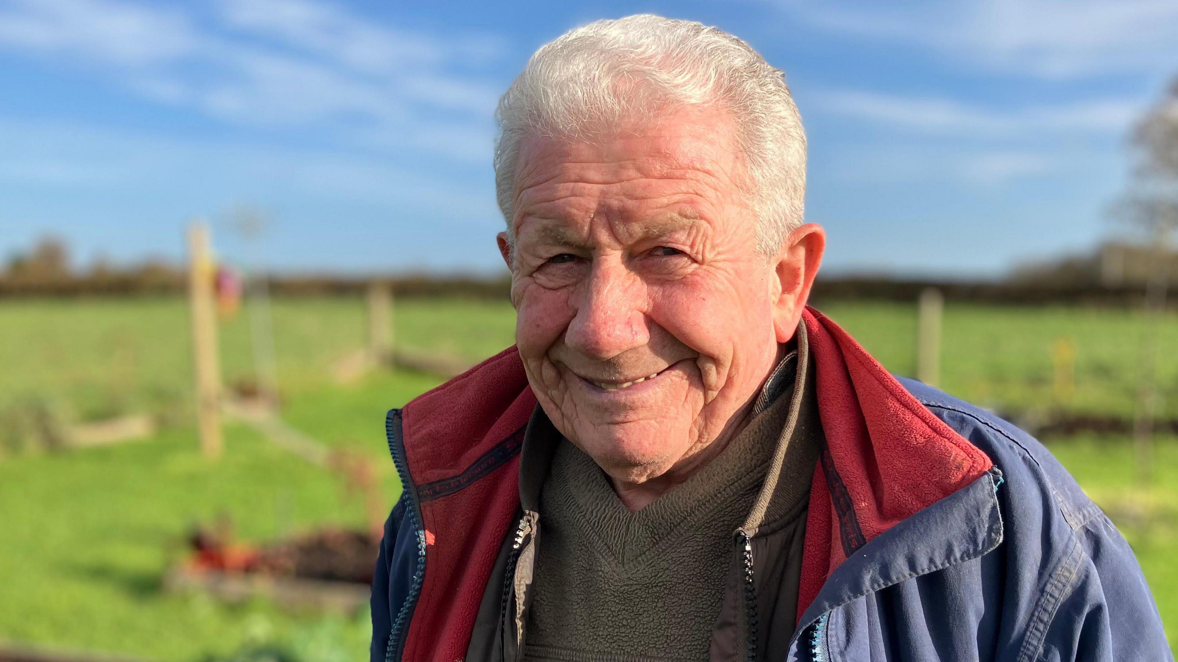 Chris in the garden. He has short white hair and is wearing a blue coast with red collars. The grass can be seen behind him.