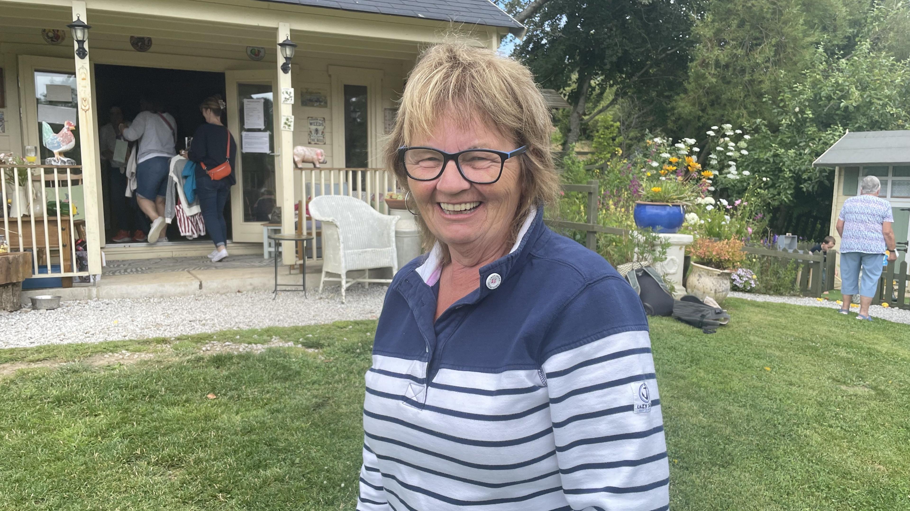 A woman with short hair and glasses and a striped blue and white top grins for the camera in front of a wooden chalet.