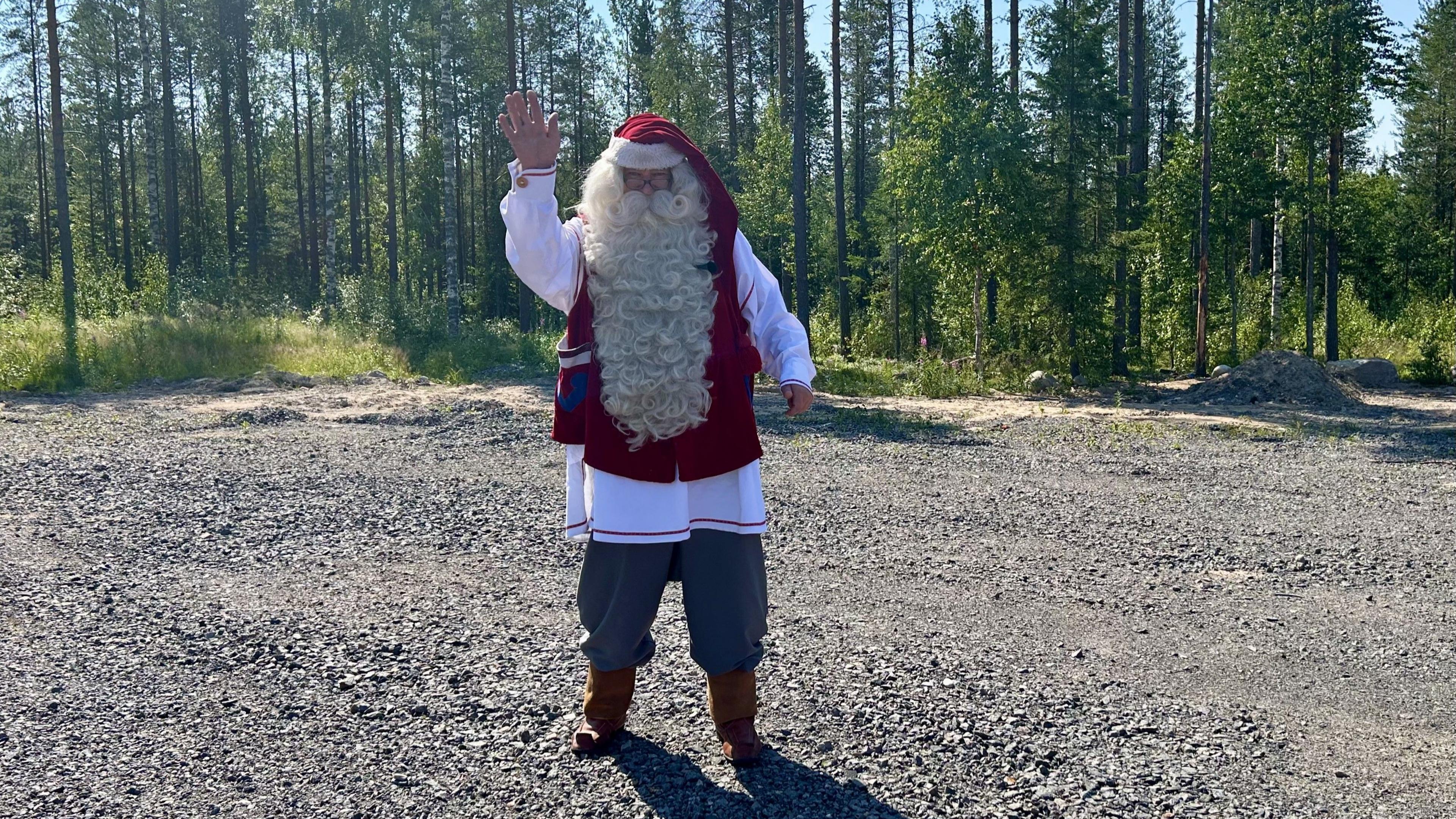 A man dressed as Santa waves at the camera. He is standing on concrete and surrounded by pine trees but the landscape in snowless
