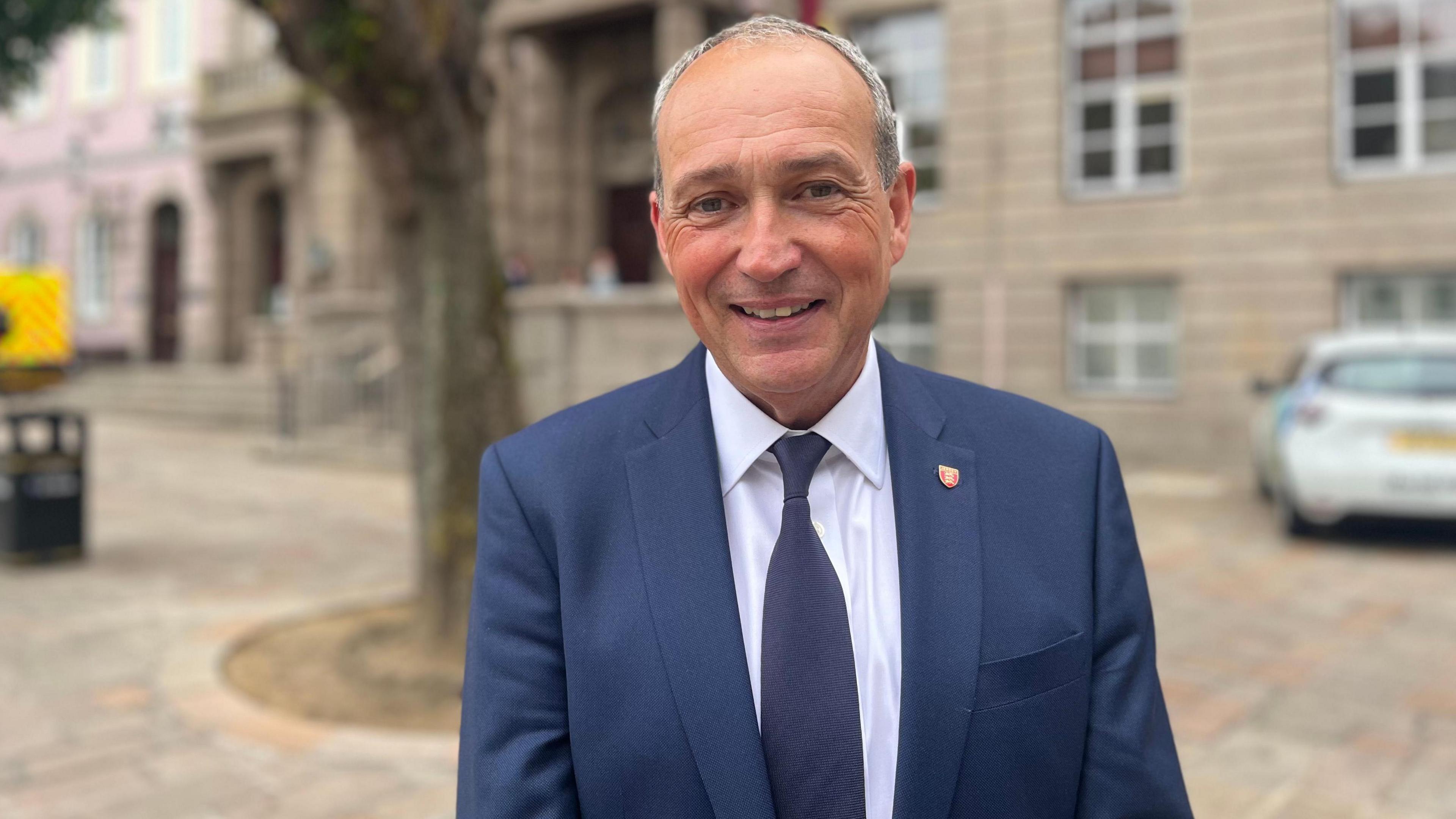 Lyndon Farnham is smiling at the camera. He is wearing a blue suit jacket with a blue tie and white shirt and is standing outside the stone-clad Royal Court in Jersey.