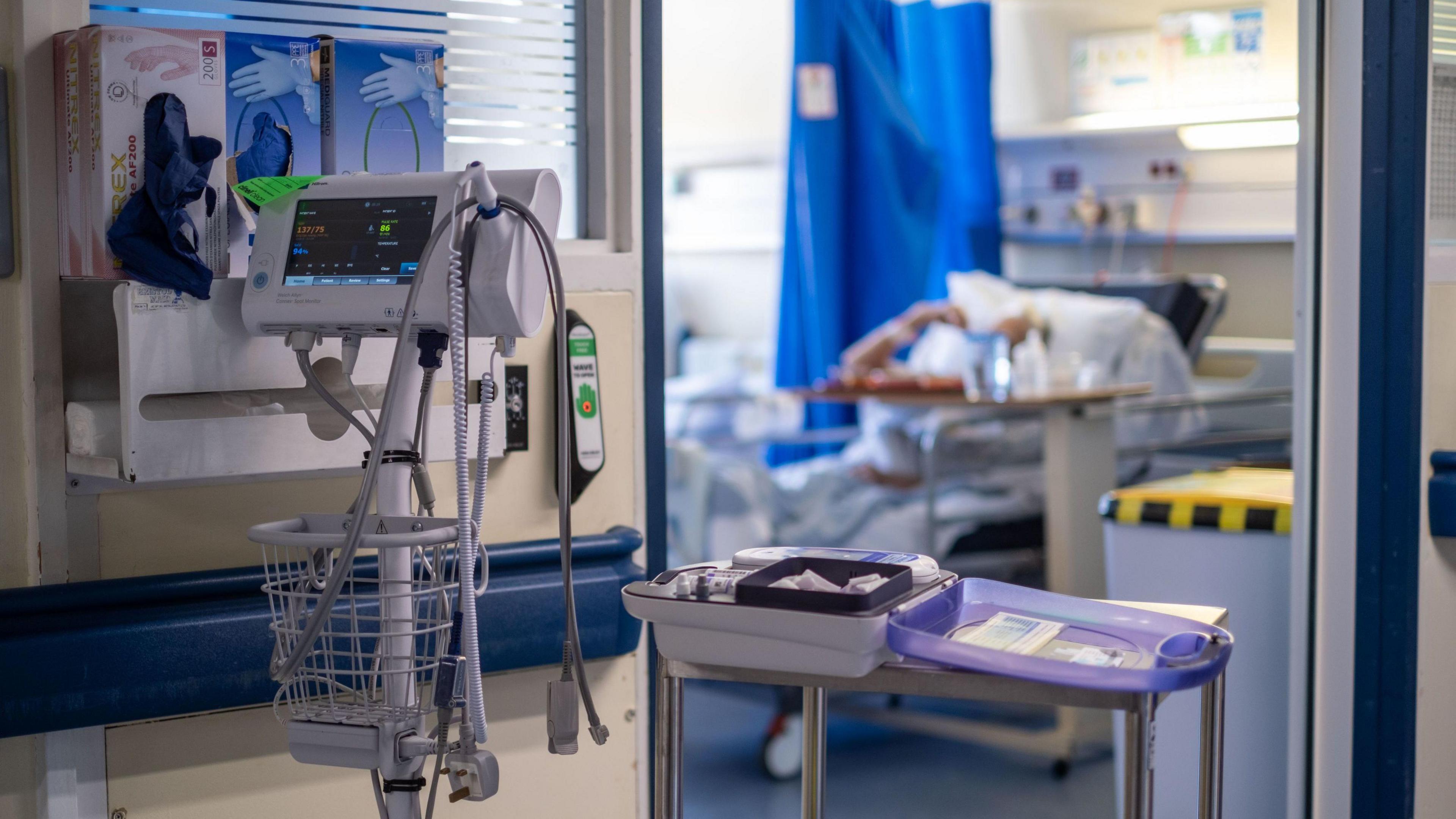 An electronic heart monitor on a trolley stands in the hallway of a hospital ward. On the wall is a dispenser for surgical gloves. Through a door, and out of focus, is a person laying on a hospital bed.