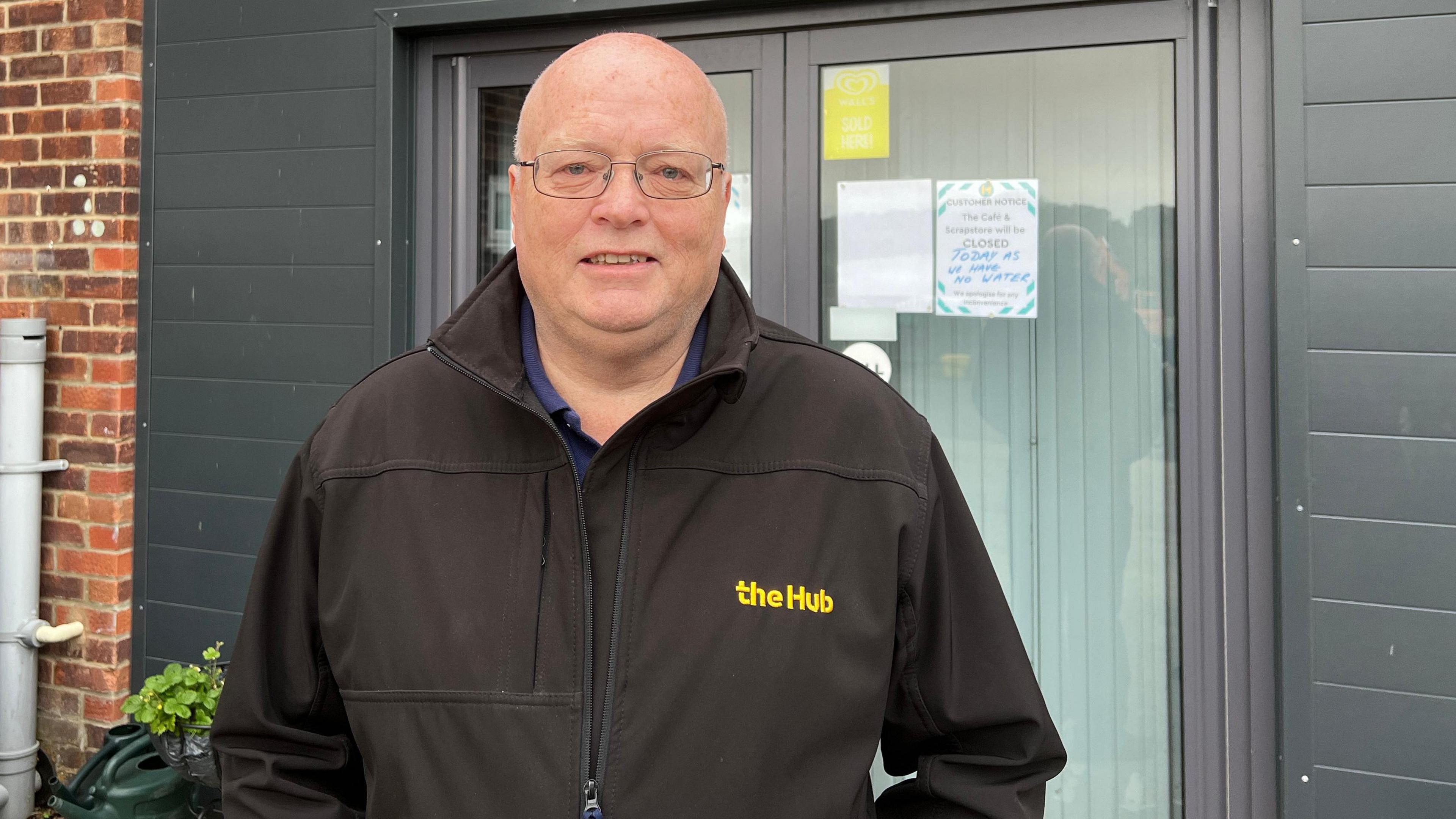 Ken Coombes wearing a black zip up jacket with 'The Hub' written in yellow font. He is bald, clean shaven, wearing thin framed glasses and standing with his hands in his pockets looking at the camera.