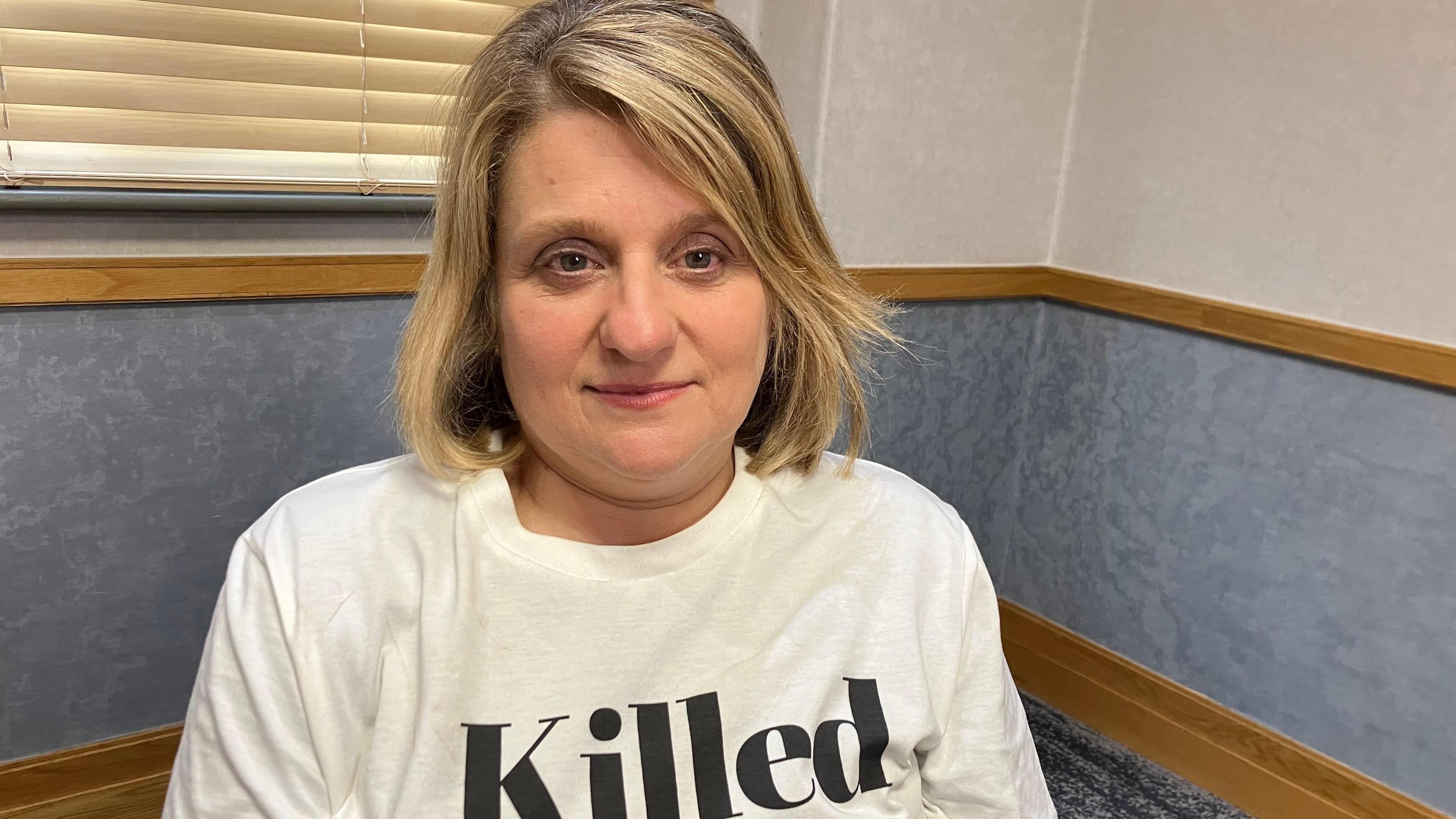 Carole with short blonde hair smiling at the camera wearing a white t shirt that says Killed Women