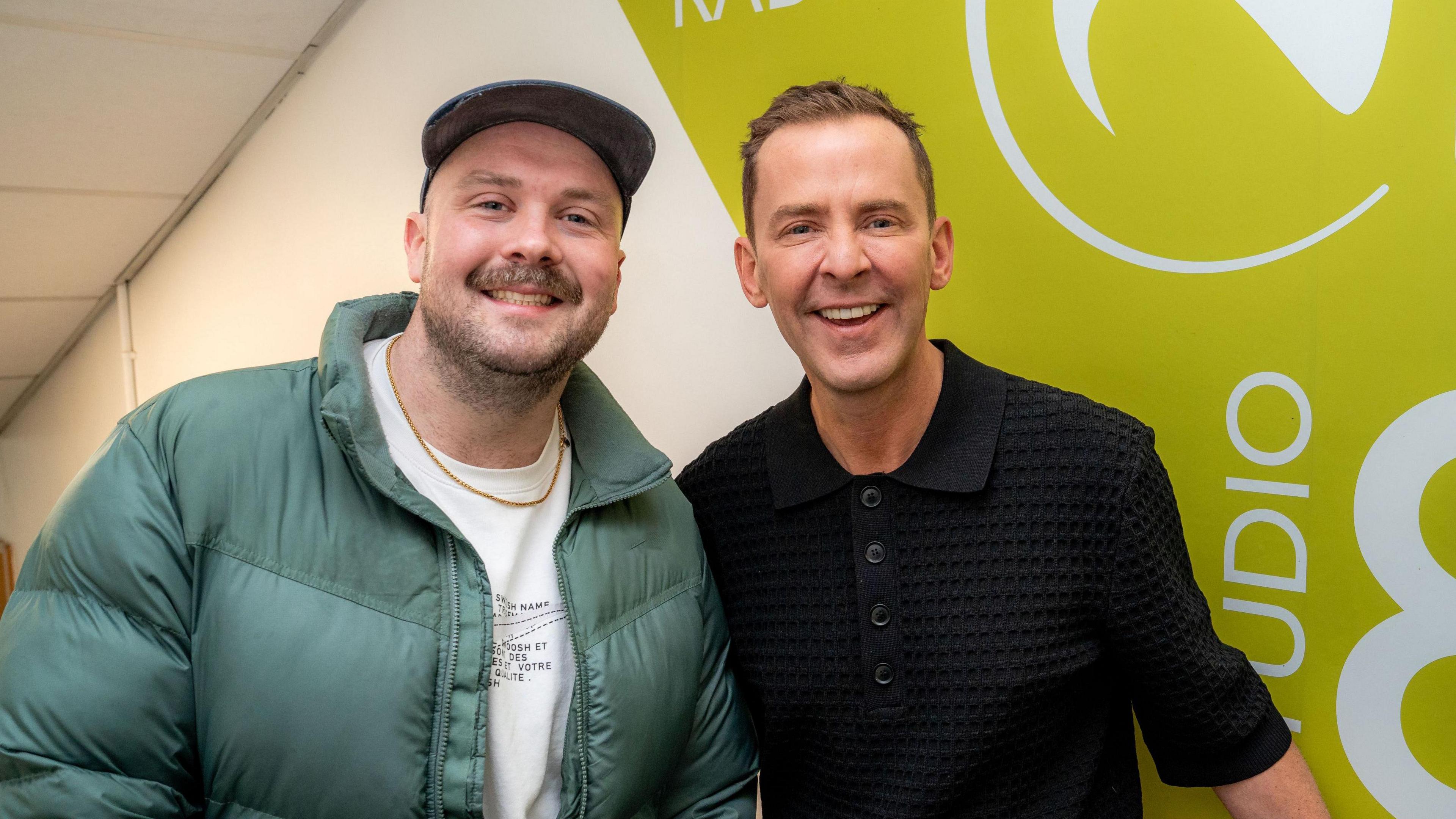 John Kerr is wearing a green puffa jacket and cap and is smiling. Scott Mills is wearing a black top with buttons up the top. 