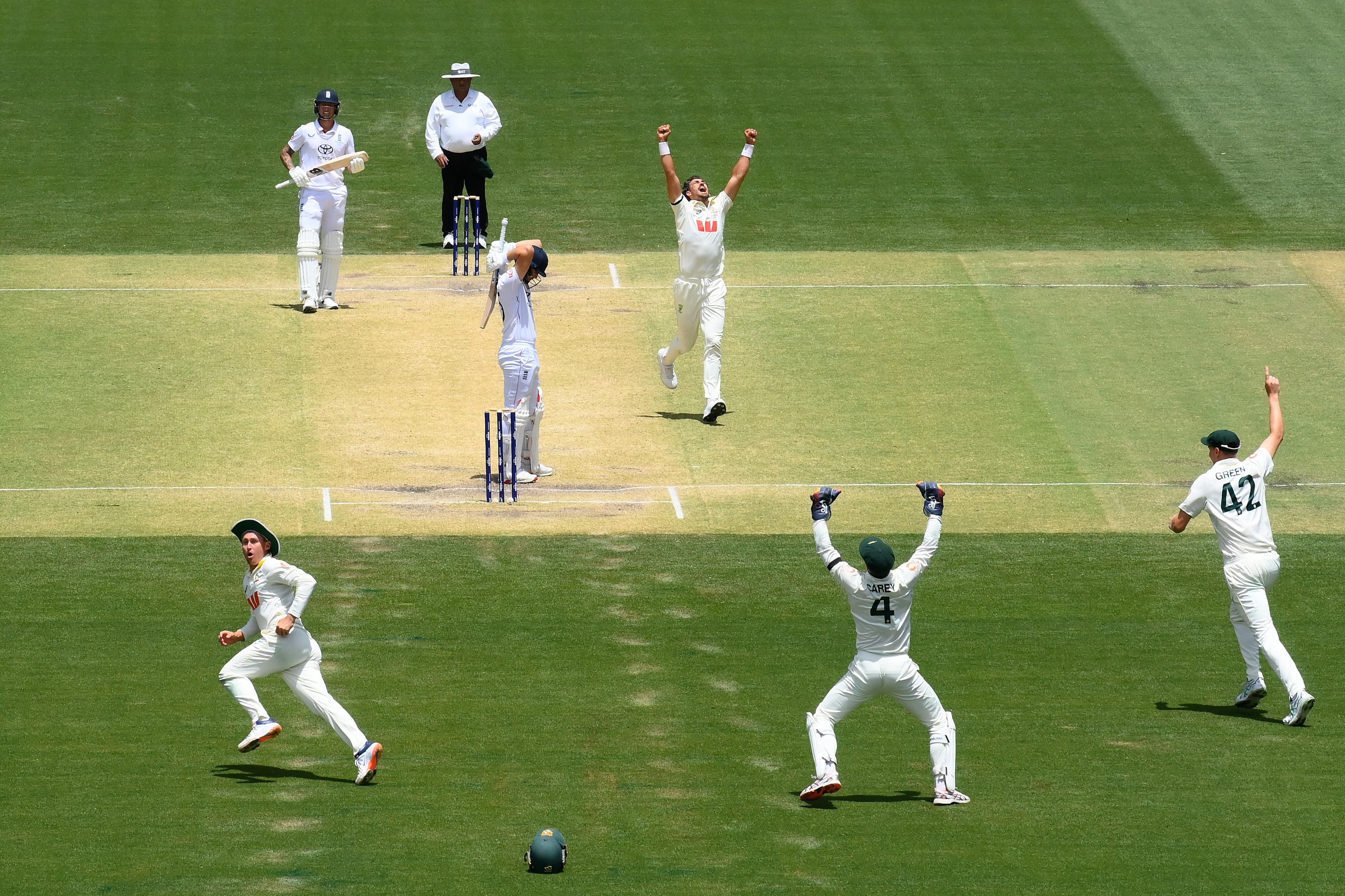 A cricket match scene showing a bowler and several fielders celebrating a dismissal, with arms raised in excitement. The batter stands near the stumps, holding their head in disappointment. The umpire watches from behind the play, and players in white uniforms are spread across the field on a bright, sunny day.
