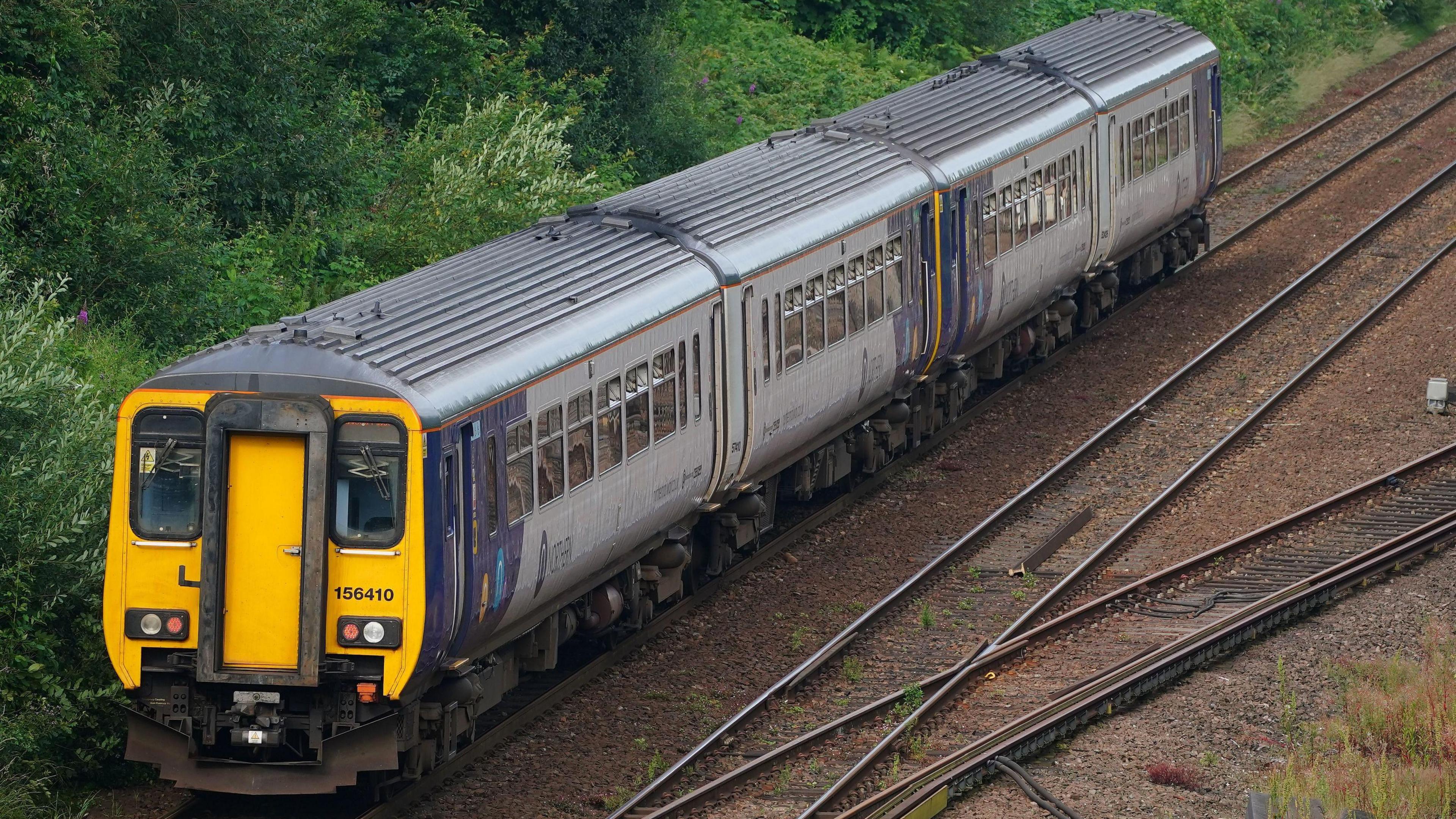 A Northern rail train on a track backed by green shrubs and bushes