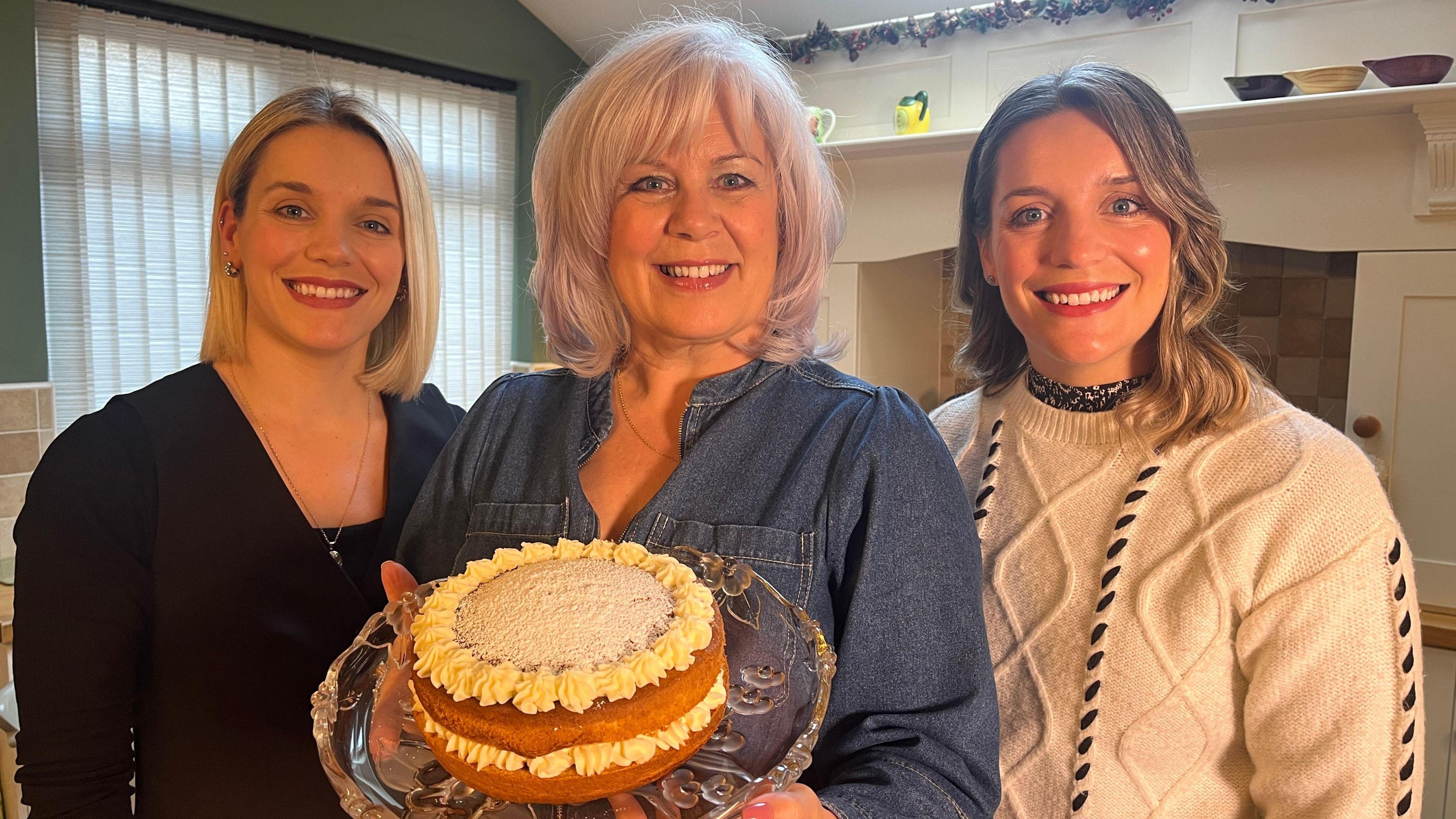 Diane Styche holds a iced sponge cake on a glass platter and is flanked by her daughters. Diane has shoulder-length grey hair and wears a blue denim shirt. To her right, Bethany has shoulder-length blonde hair and wears a black top. To her left, Charlotte has shoulder-length brown hair and wears a white woollen jumper.