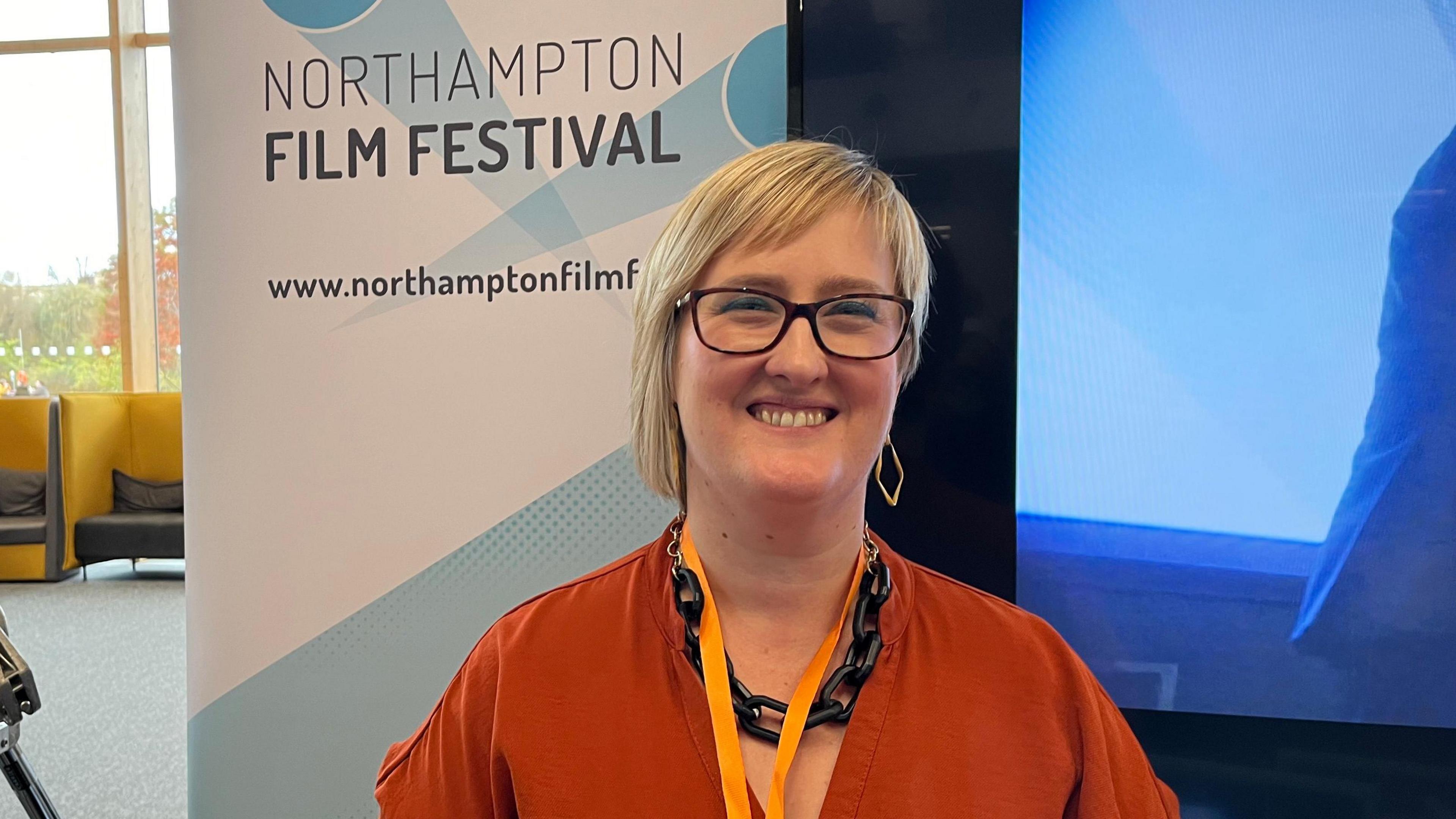 Becky Carrier with medium-length blond hair wearing an orange top and lanyard. A sign behind her says "Northampton Film Festival".