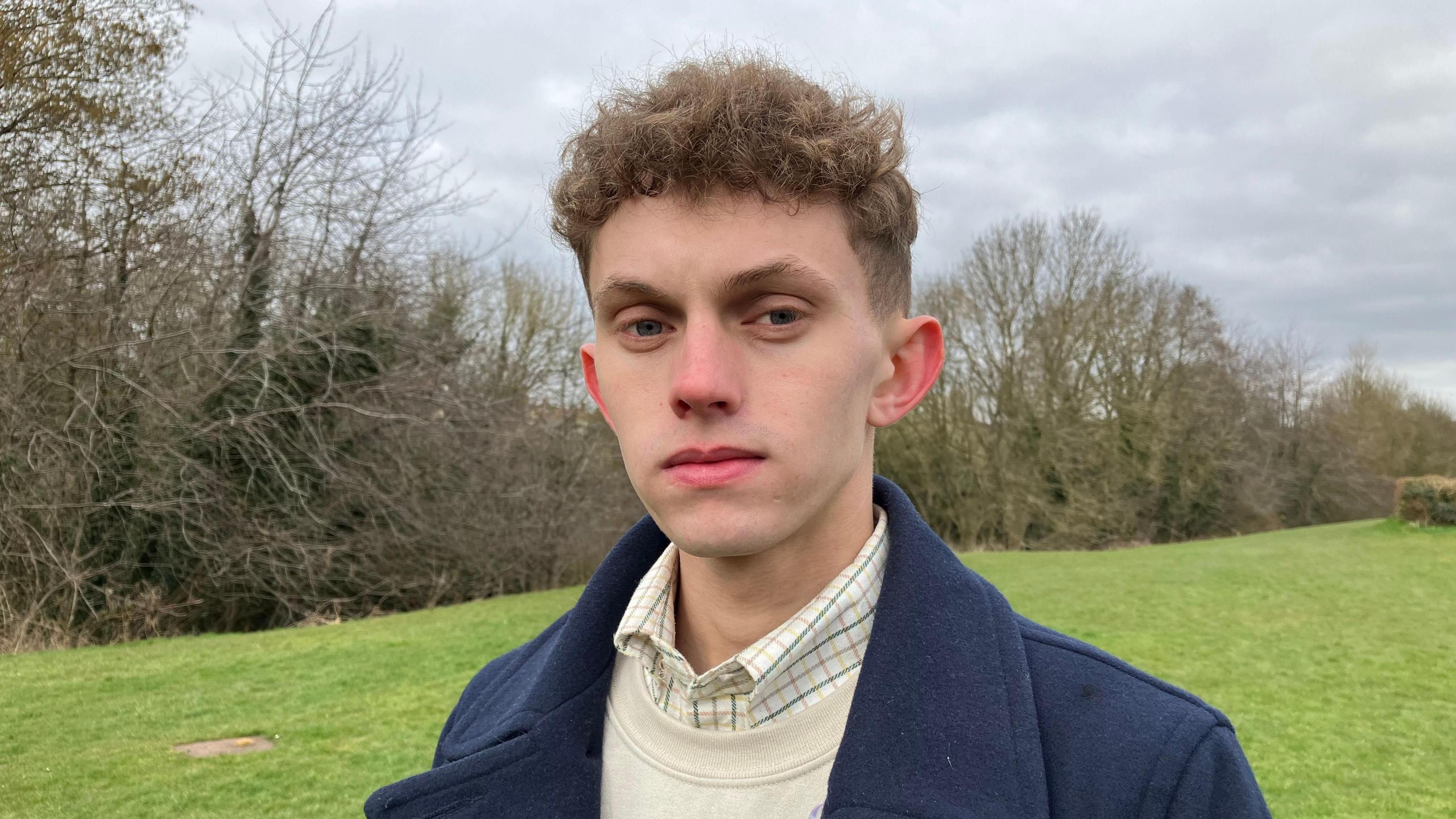 Man stares at the camera with a blank expression. He is stood in a field with bare branches lining a hedgerow. He wears a checked shirt, dram sweatshirt and thick navy blue coat.