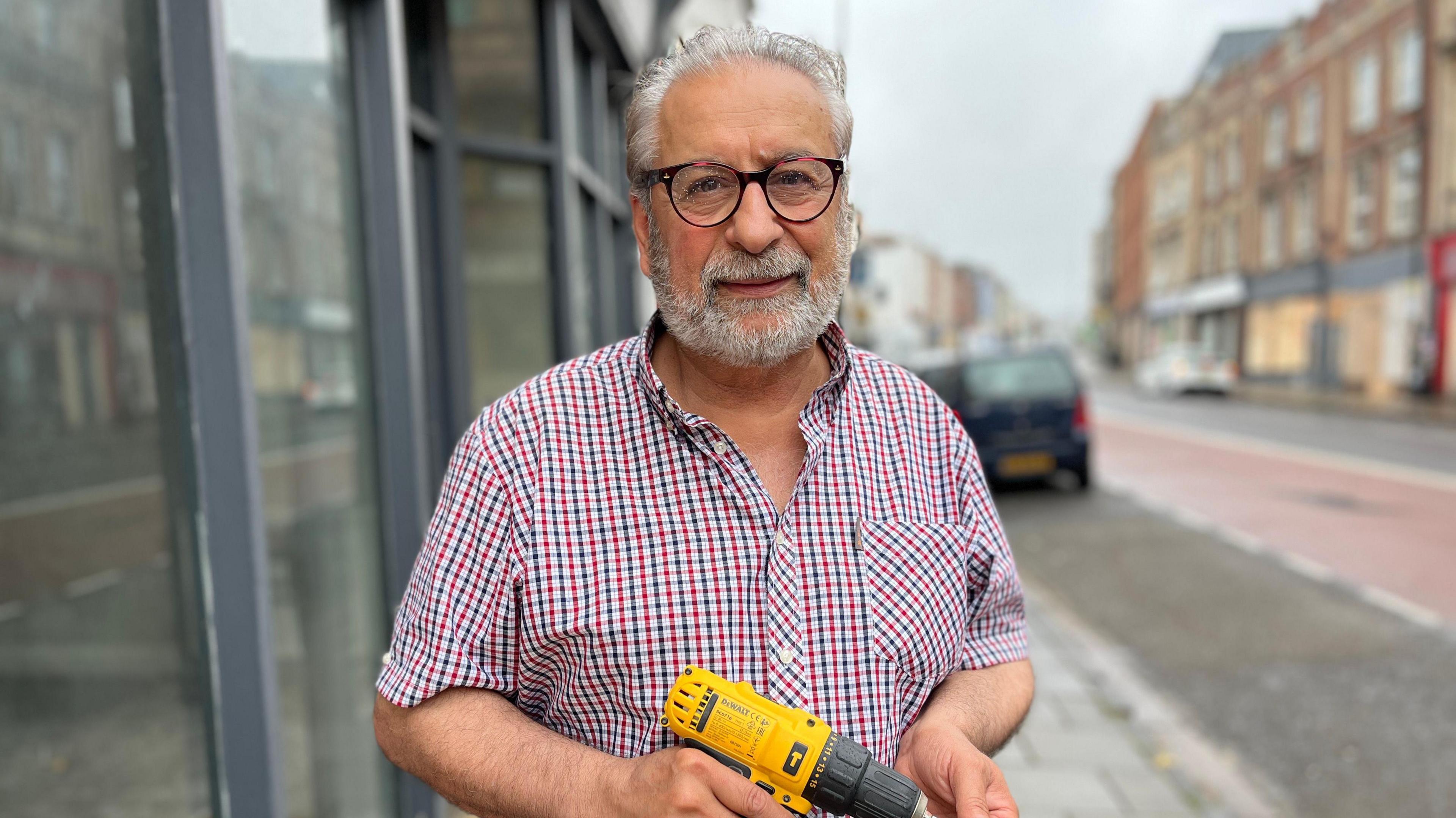 Essie Golshan standing outside his shop on Old Market, with the street visible behind him. Some buildings still have boards over the windows. He has grey hair and wears round dark glasses and a blue, red and white checked shirt. He holds a yellow and grey drill. 