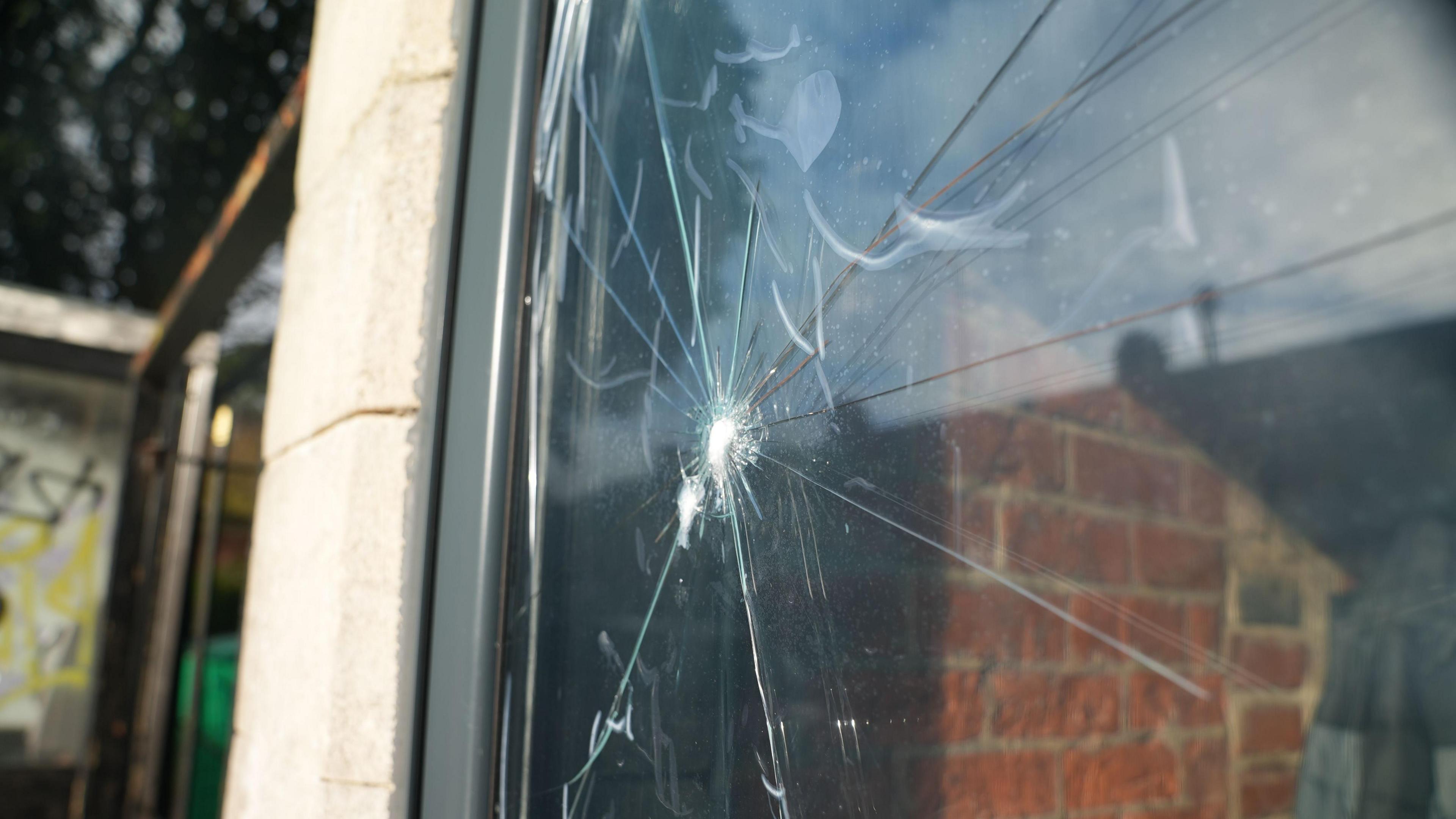 Close up photograph of a smashed window. There is clear film over the top of the crack. Red brick wall can be seen through the window to the inside of the coffee shop. 