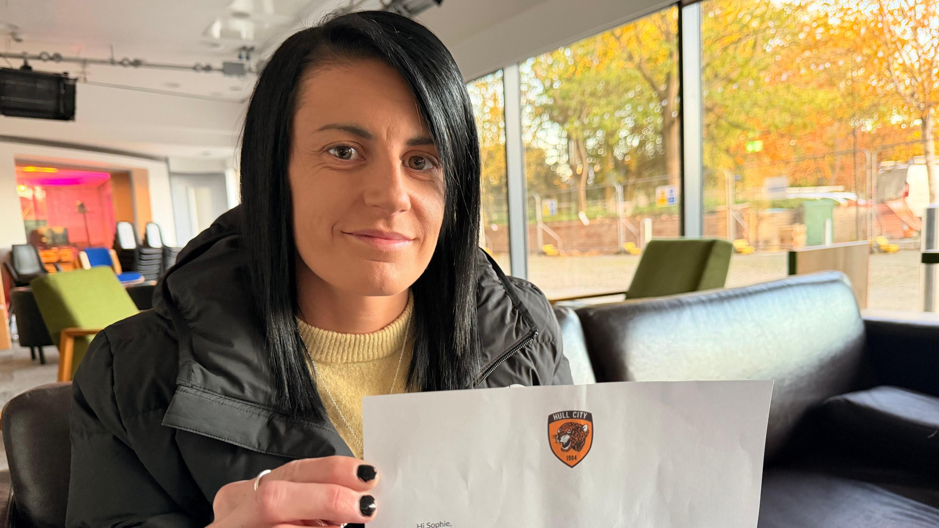 Sophie Griffiths looking at the camera holding a letter with a Hull City logo at the top. She has long black straight hair with a side parting. She is wearing a black coat and a yellow sweater. Her nails are painted black. She is sitting on a black sofa inside an office-like space.